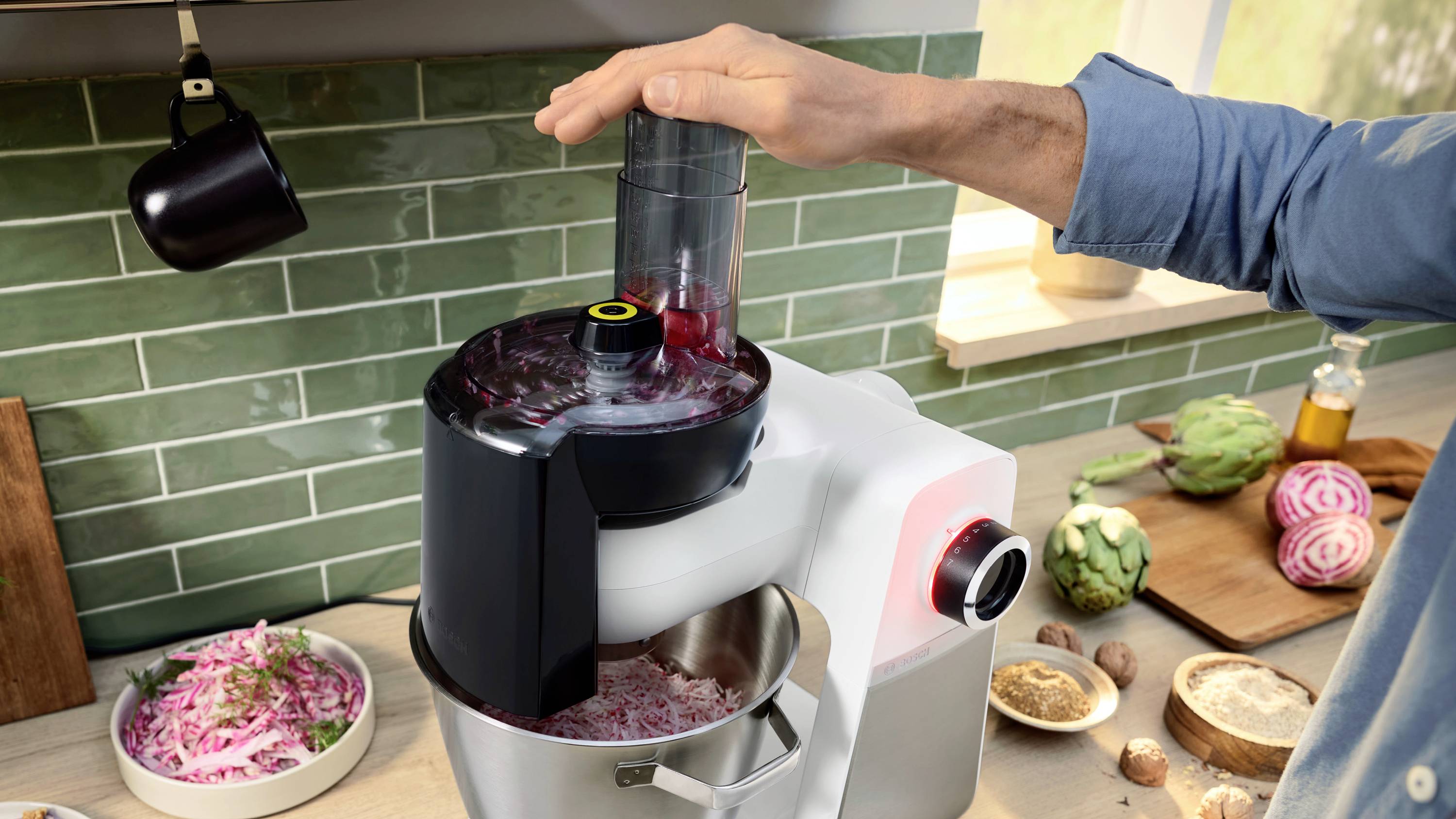 Eine Person bedient einen Küchenroboter mit Zwiebeln darin, auf einer Küchenarbeitsplatte mit Gemüse und Gewürzen, was auf einen laufenden Kochvorgang hinweist.