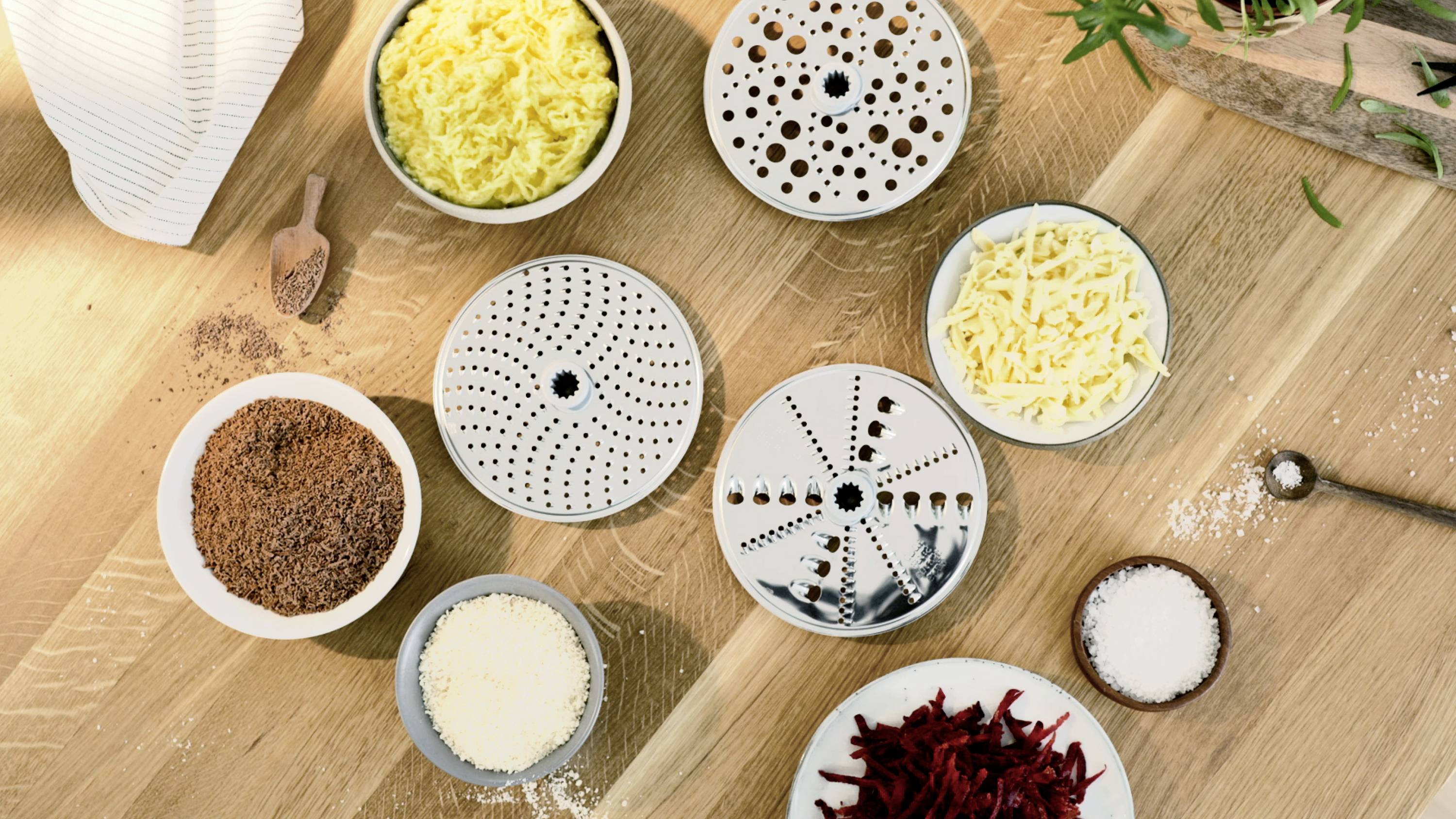 Käse, Schokolade und Rote Bete liegen in Schalen neben Reiberaufsätzen auf einer Holzoberfläche, bereit zur Lebensmittelzubereitung.