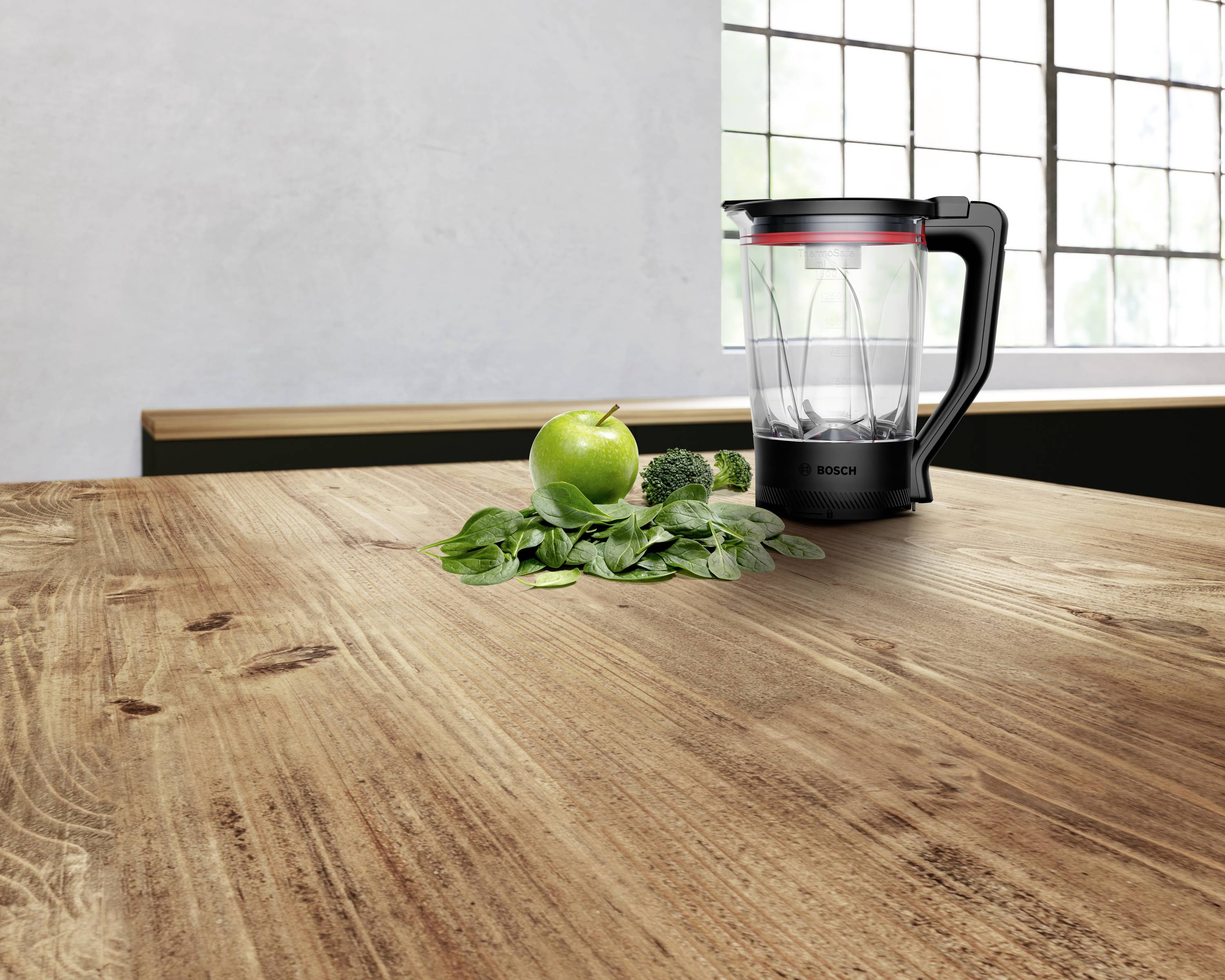 Ein Mixer mit einem durchsichtigen Behälter steht auf einem Holztisch neben Spinat, einem Apfel und Brokkoli, was die Vorbereitung eines Smoothies andeutet.