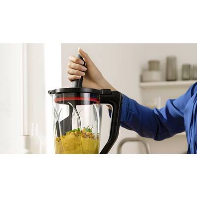 Eine Person in einem blauen Hemd verwendet einen Standmixer mit schwarzem Griff, um Kichererbsen und Kräuter zu mischen und in einer Küchenumgebung ein cremiges Püree zuzubereiten.