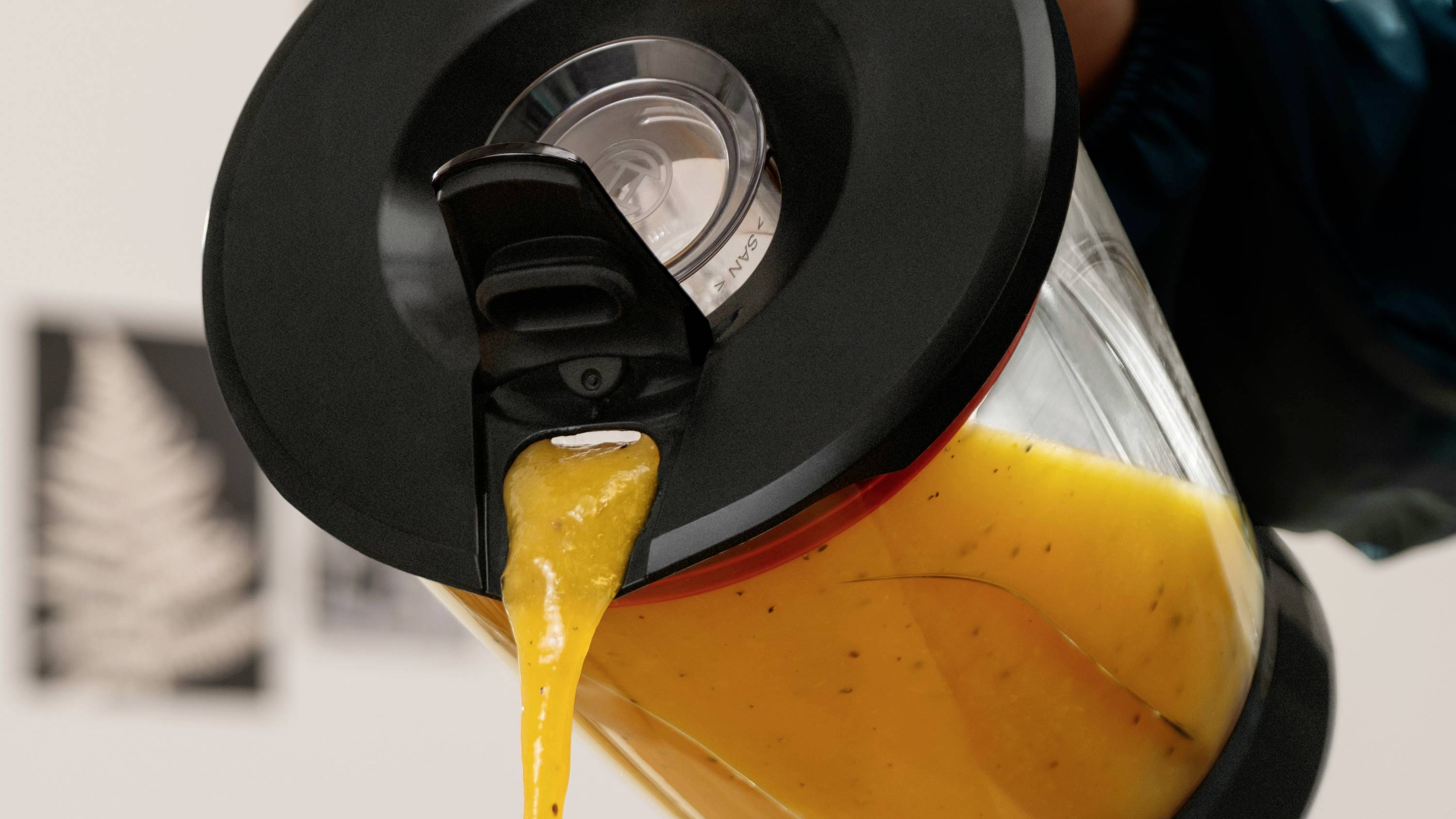 Eine Hand gießt eine dicke orangefarbene Flüssigkeit aus einem schwarzgedeckelten Krug, wahrscheinlich Saft oder Smoothie, in ein Gefäß, vor einem unscharfen Hintergrund.