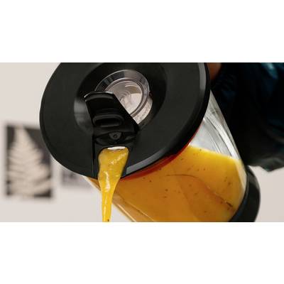 Eine Hand gießt eine dicke orangefarbene Flüssigkeit aus einem schwarzgedeckelten Krug, wahrscheinlich Saft oder Smoothie, in ein Gefäß, vor einem unscharfen Hintergrund.