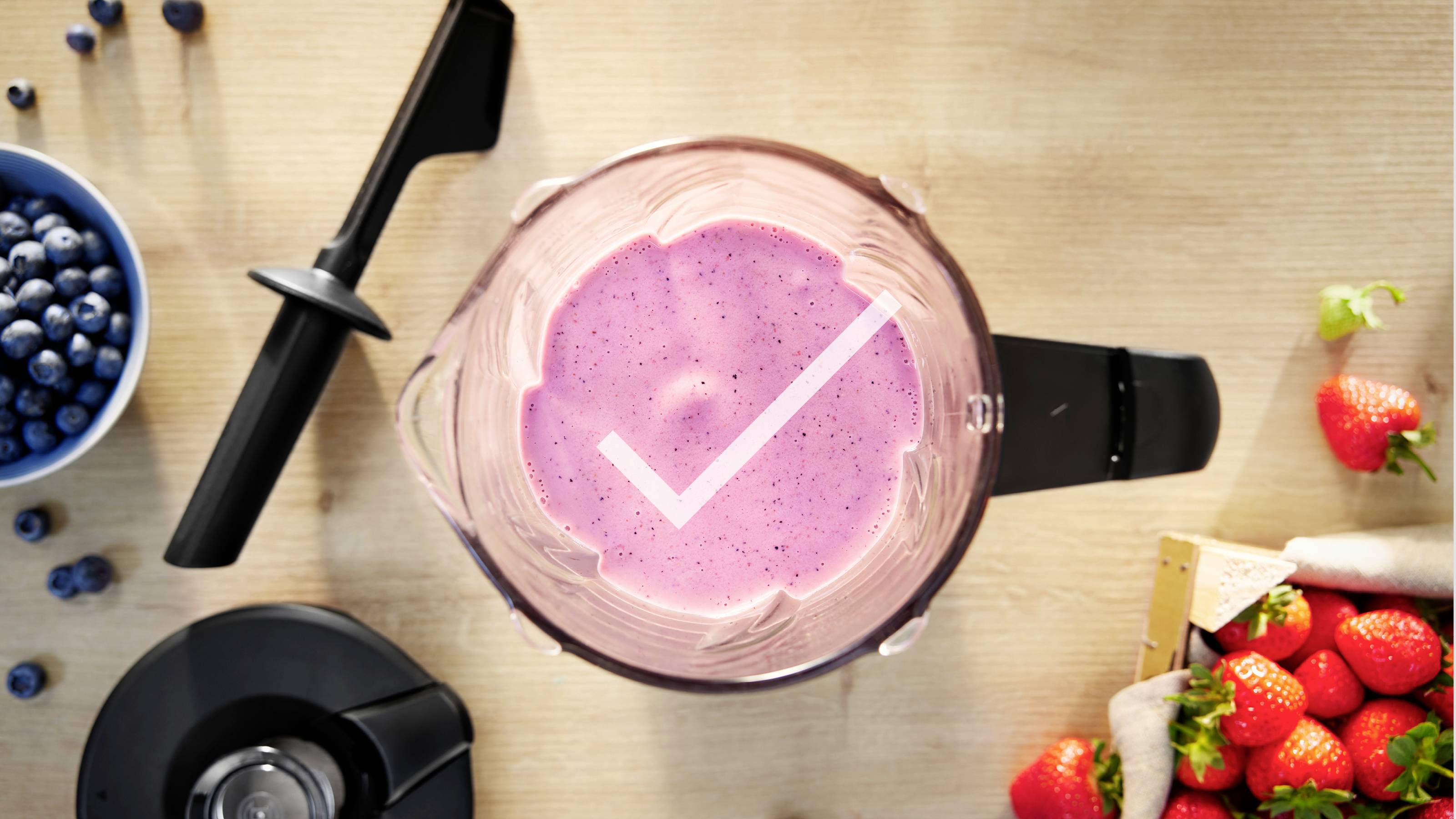 Ein Mixer mit einem rosa Smoothie im Inneren, einem Häkchen-Symbol auf der Oberfläche, umgeben von Blaubeeren, Erdbeeren und Mixerteilen.