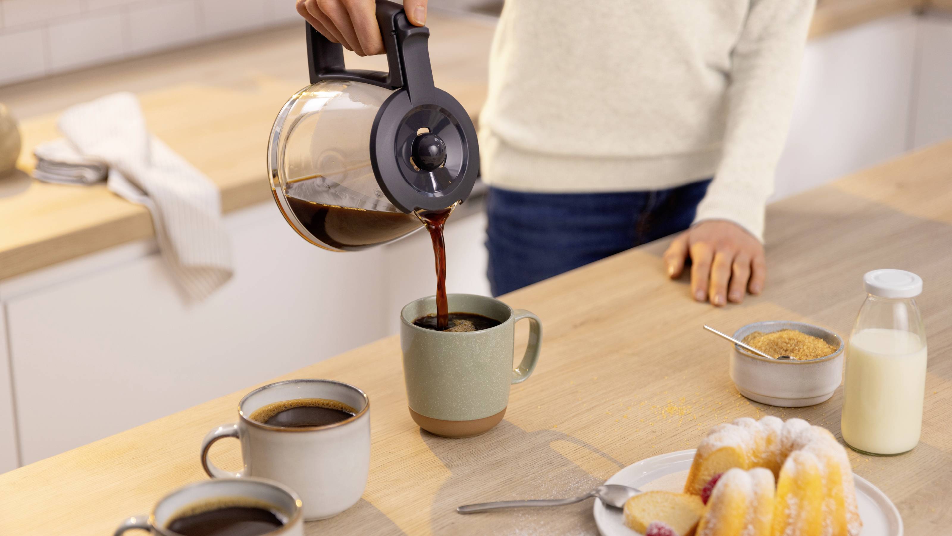 Eine Person gießt Kaffee aus einer Kanne in eine Tasse auf einer Küchenarbeitsplatte, umgeben von zwei weiteren Tassen, einem Teller mit Gebäck, Zucker und Milch.