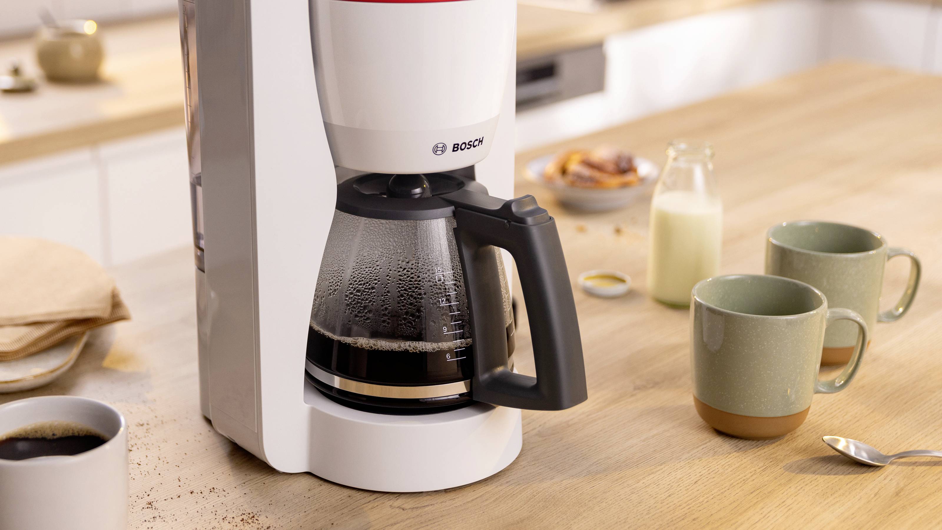 Eine Kaffeemaschine, die Kaffee brüht, mit einer halbvollen Kanne auf einer Küchenarbeitsplatte. In der Nähe zwei grüne Tassen, ein Glas Milch und eine Schüssel mit Snacks.