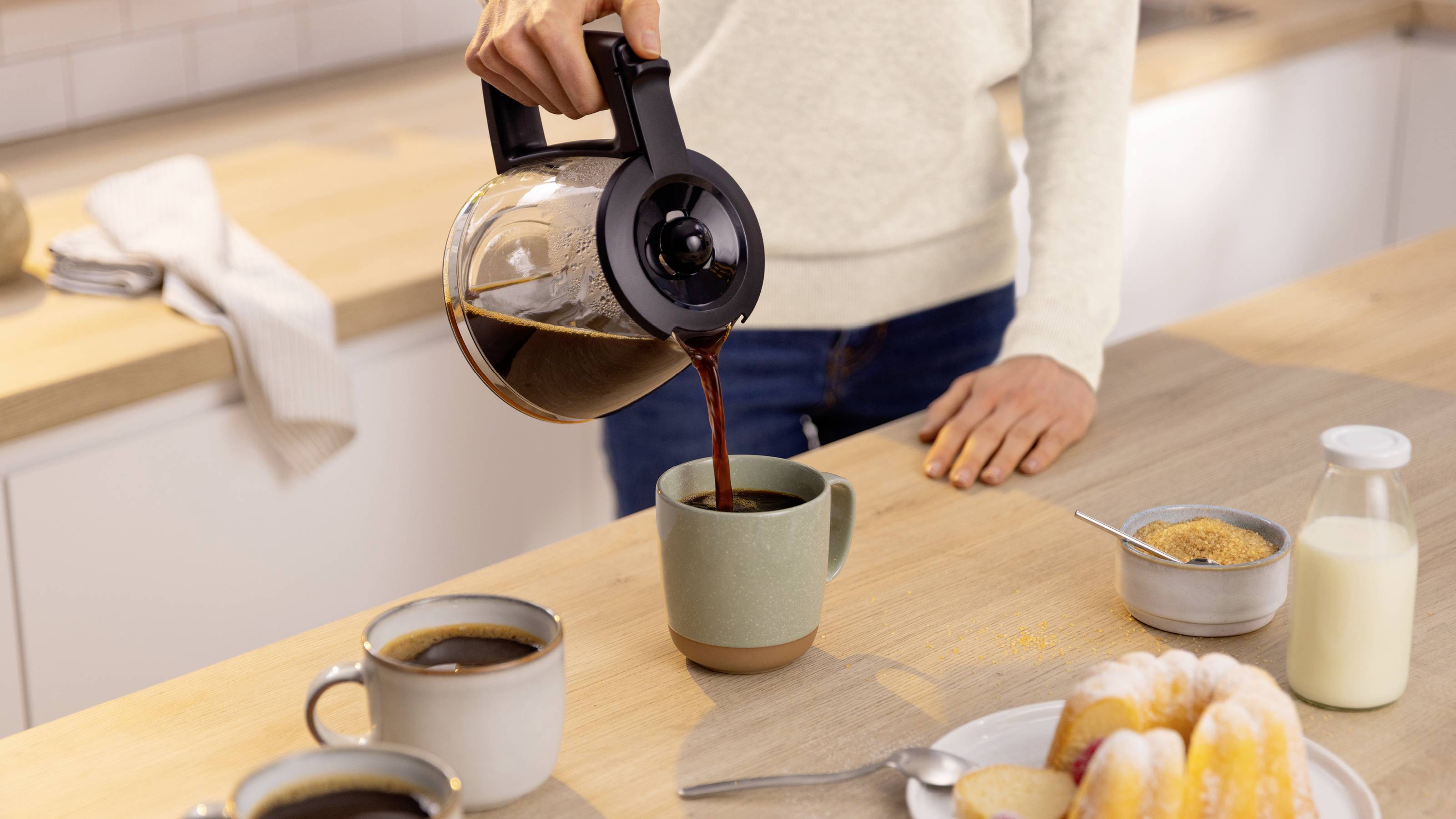 Eine Person gießt Kaffee aus einer Kanne in eine Tasse auf einer Küchenarbeitsfläche. Zwei weitere gefüllte Tassen, Zucker und Gebäckstücke befinden sich in der Nähe.