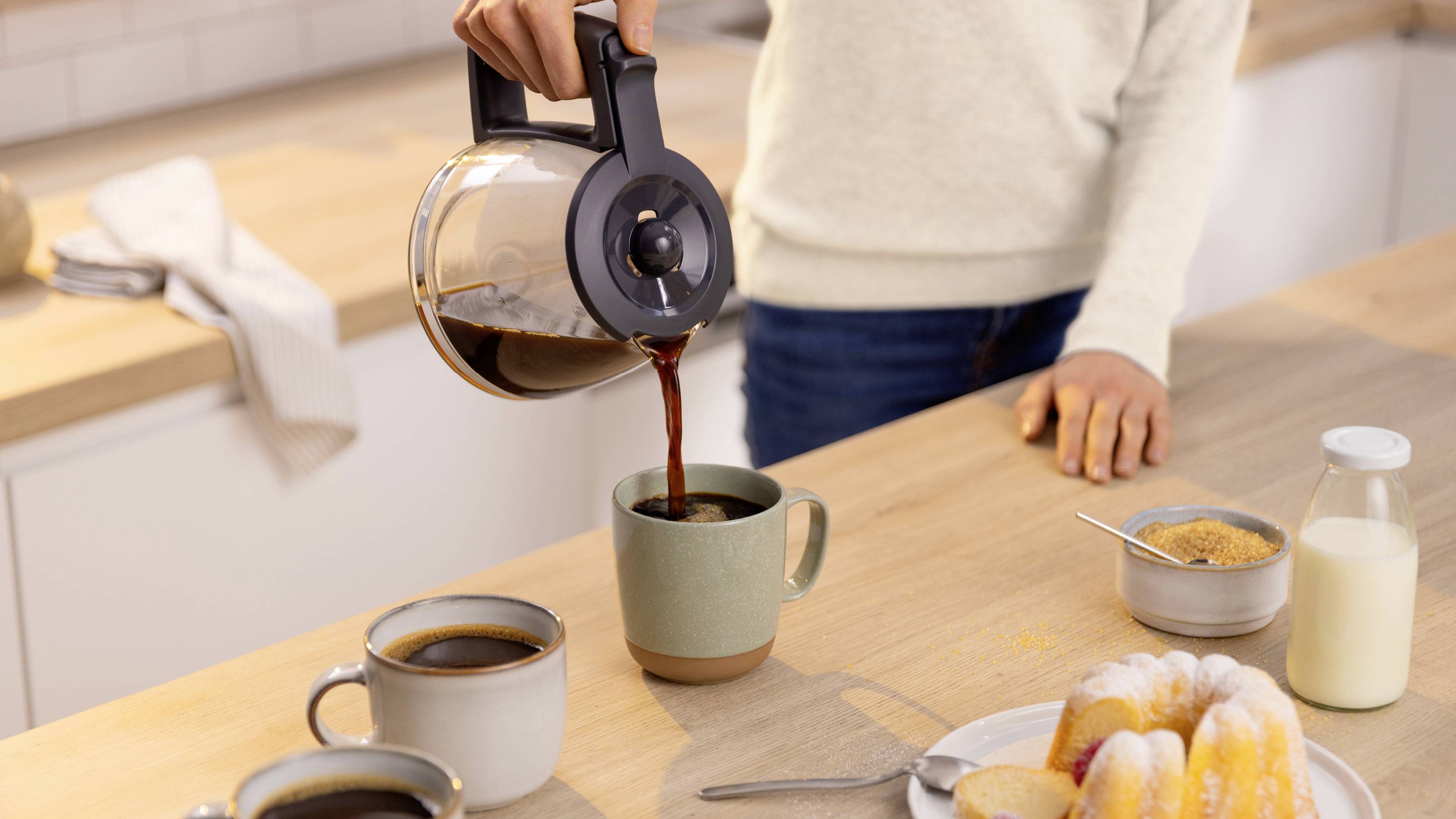 Eine Person gießt Kaffee aus einer Kanne in eine Tasse auf der Küchenarbeitsplatte, umgeben von anderen Tassen, einem Teller mit Gebäck, Milch und Zucker.