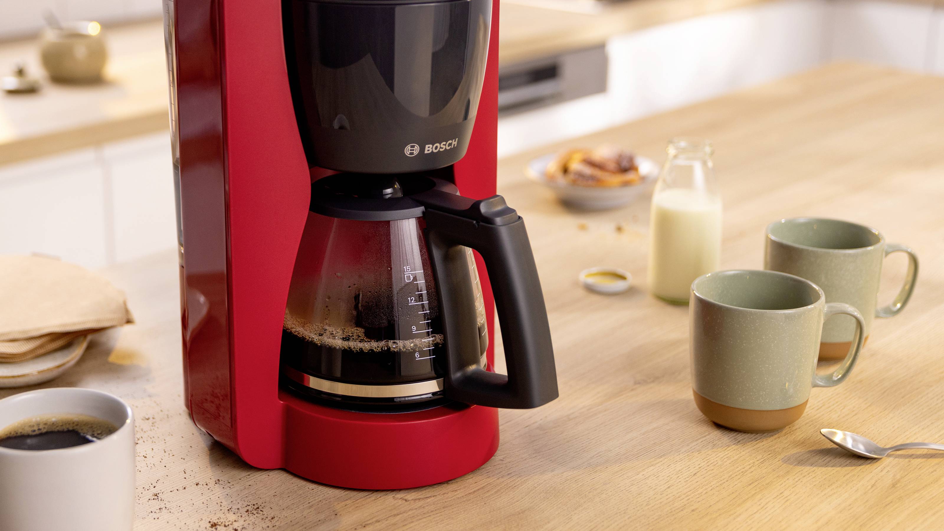 Ein roter Kaffeemaschine mit einer vollen Glaskanne steht auf einer Holzarbeitsplatte, umgeben von zwei Tassen, einer Milchflasche und Kaffeebohnen.