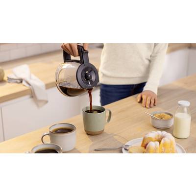 Eine Person gießt Kaffee in eine Tasse auf einer Küchenarbeitsplatte. In der Nähe stehen zwei gefüllte Tassen, ein Teller mit Gebäck, ein Zuckerglas und eine Milchflasche.