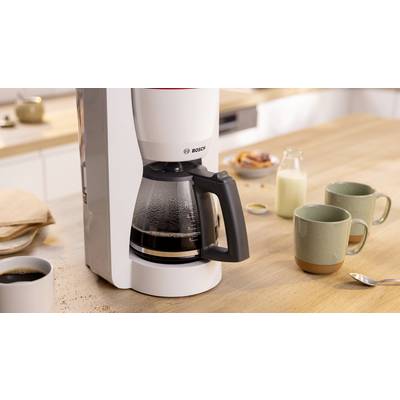 Eine weiße Kaffeemaschine mit einer Glaskanne, gefüllt mit gebrühtem Kaffee, steht auf einer Holztheke. In der Nähe befinden sich zwei Tassen und eine Milchflasche.