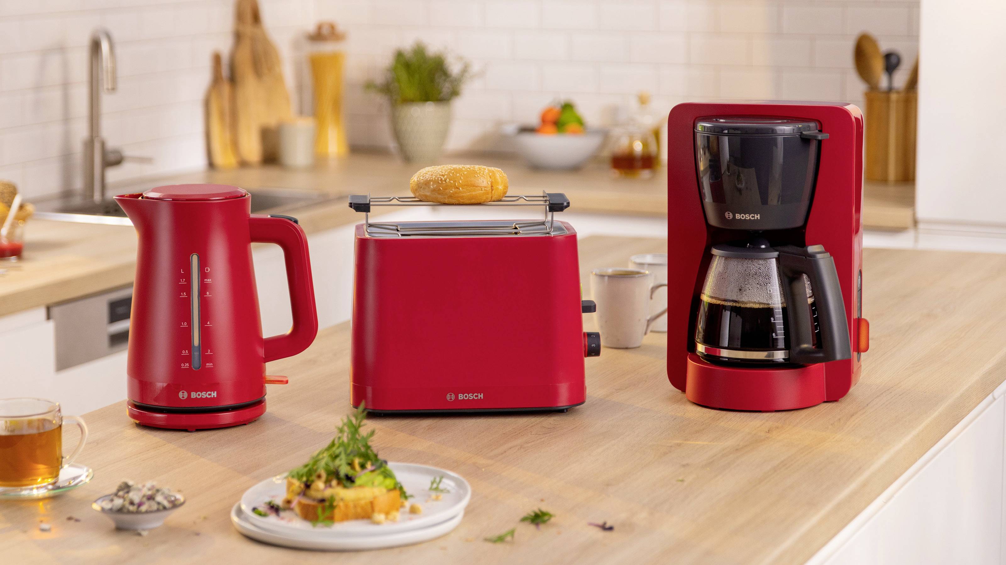 Ein roter Küchengeräte-Satz mit einem elektrischen Wasserkocher, einem Toaster mit Brot und einer Kaffeemaschine auf einer Holzarbeitsplatte mit Lebensmitteln und Getränken.