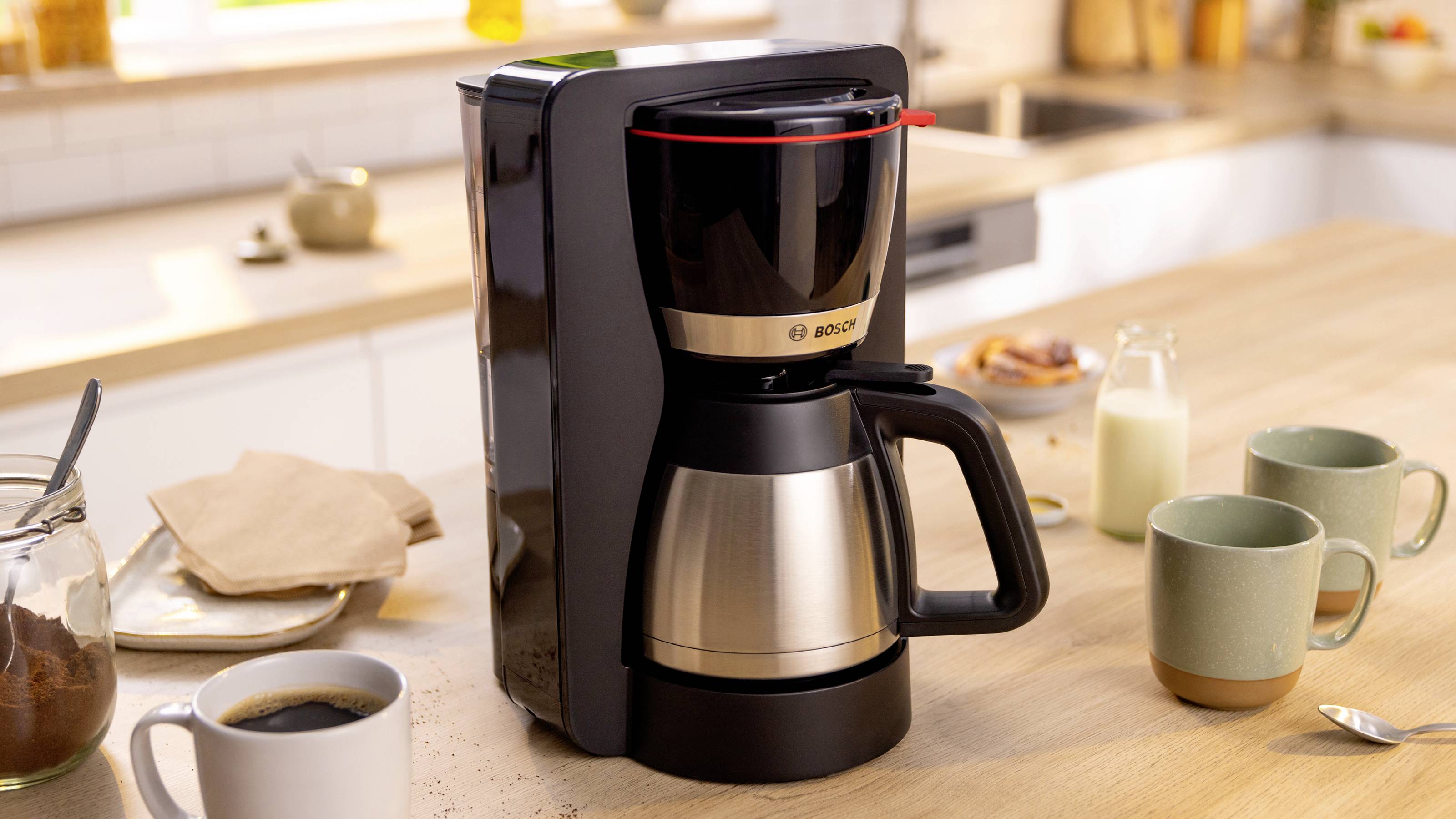 Eine Kaffeemaschine auf einer Küchenarbeitsplatte, umgeben von Tassen, einem Glas Kaffee, einem Krug Milch und einer Zuckerschale, was eine morgendliche Kaffeevorbreitung andeutet.