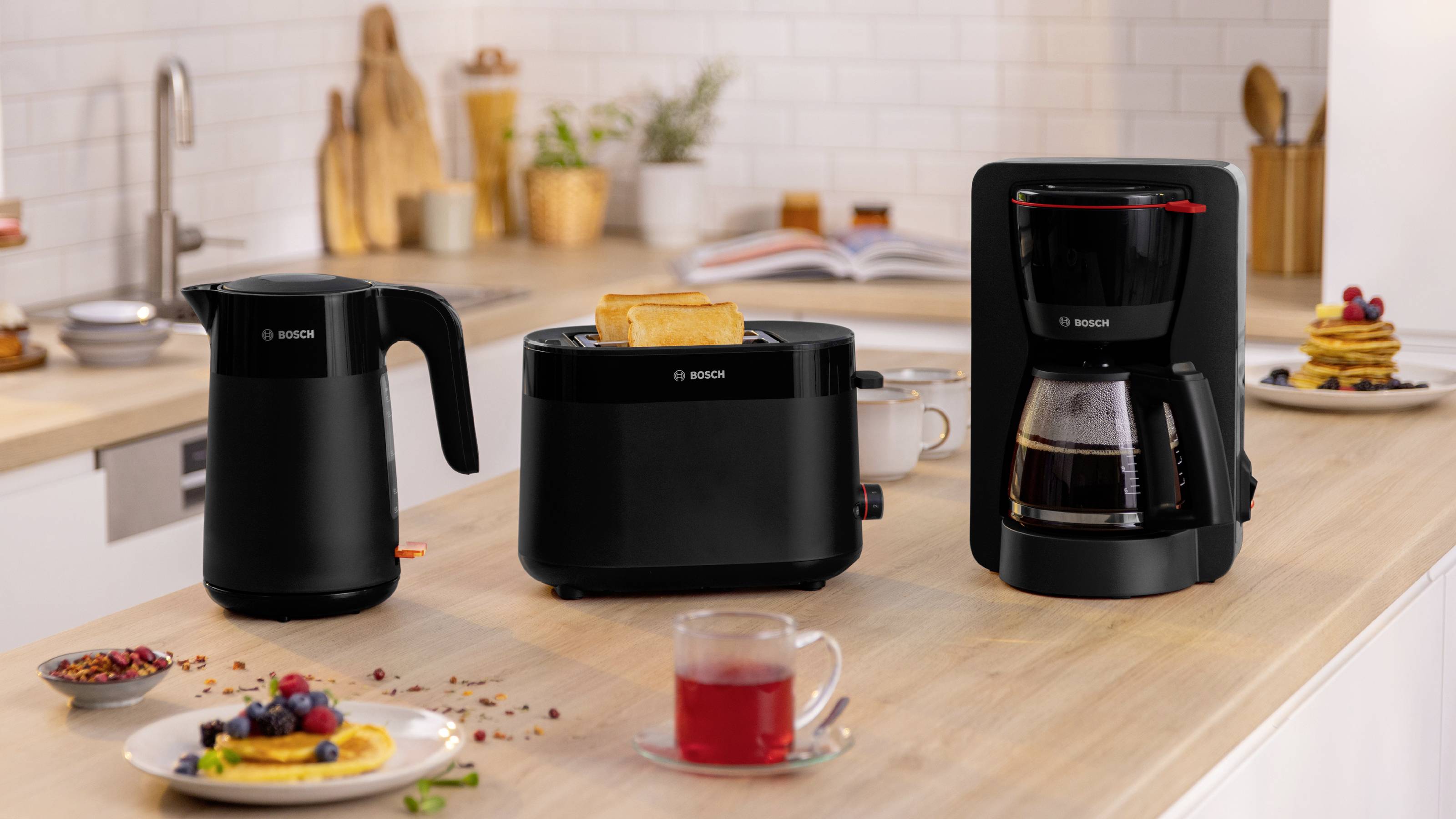 Eine moderne Küchenarbeitsplatte mit einem schwarzen Wasserkocher, einem Toaster mit Toast und einer Kaffeemaschine. Im Hintergrund sind Pfannkuchen und Früchte zu sehen.