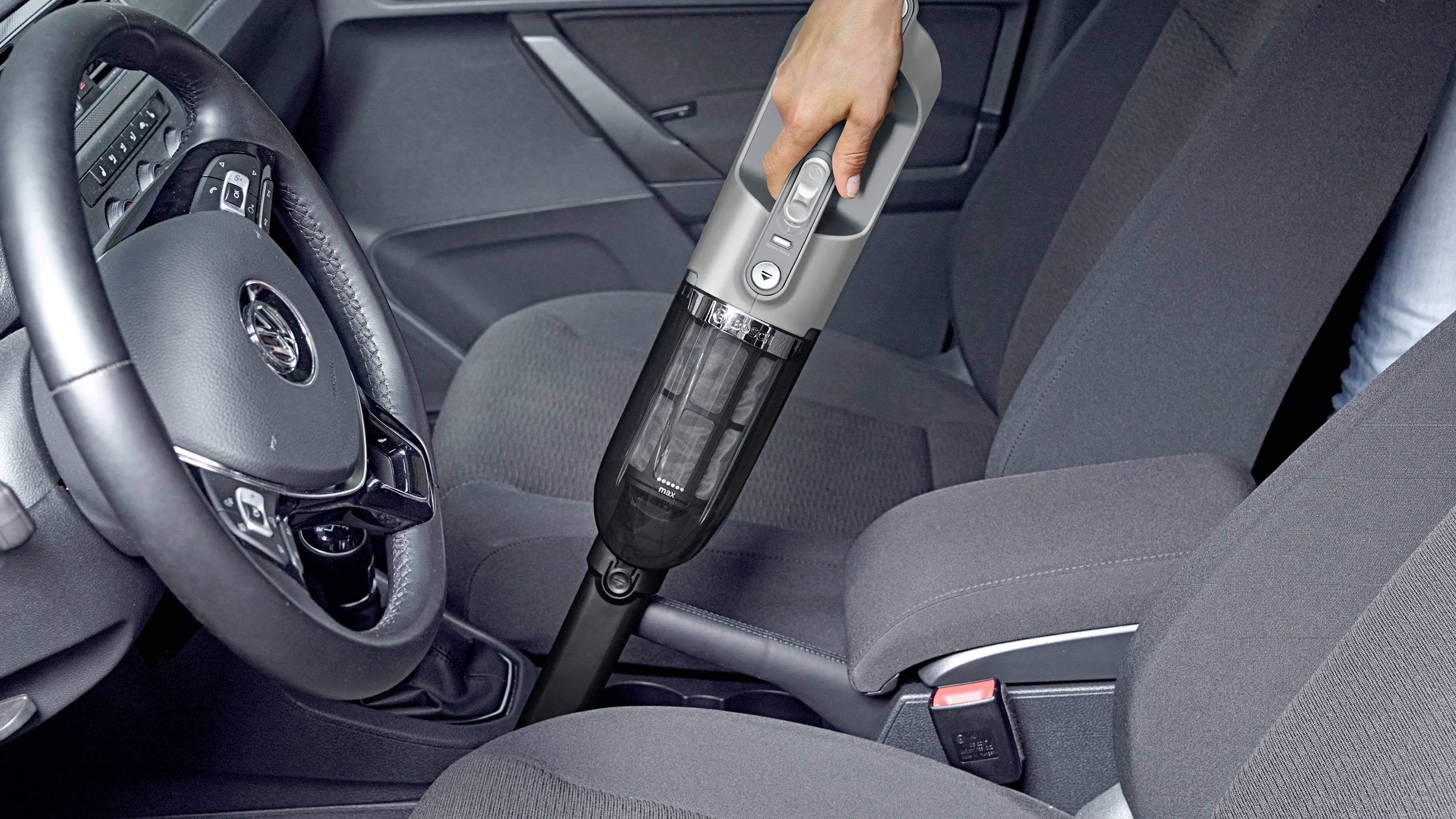 Eine Person benutzt einen tragbaren Staubsauger, um das Wageninnere zu reinigen und konzentriert sich dabei auf den Bereich zwischen Fahrer- und Beifahrersitz.