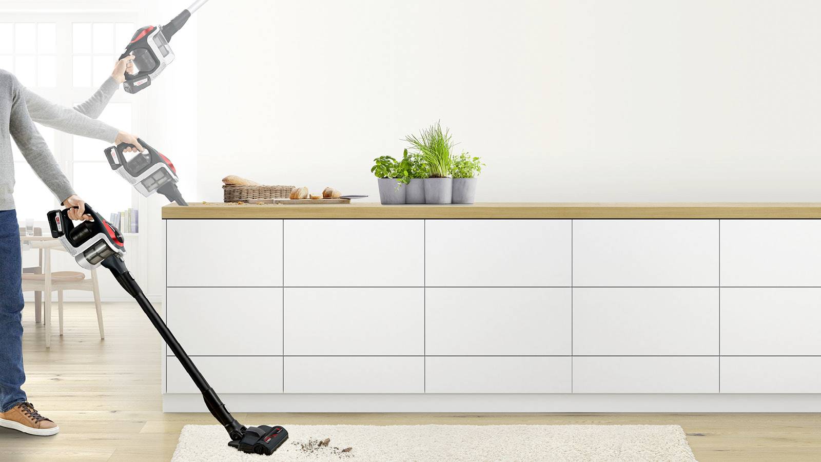 Eine Person benutzt einen kabellosen Staubsauger auf einem weißen Teppich in einer modernen Küche und zeigt die flexible Handhabung des Staubsaugers in verschiedenen Positionen.