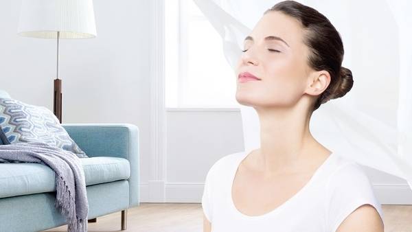 Eine Frau in einem weißen Hemd sitzt friedlich, die Augen geschlossen, in einem hellen Raum mit einer Couch und einer Lampe im Hintergrund.