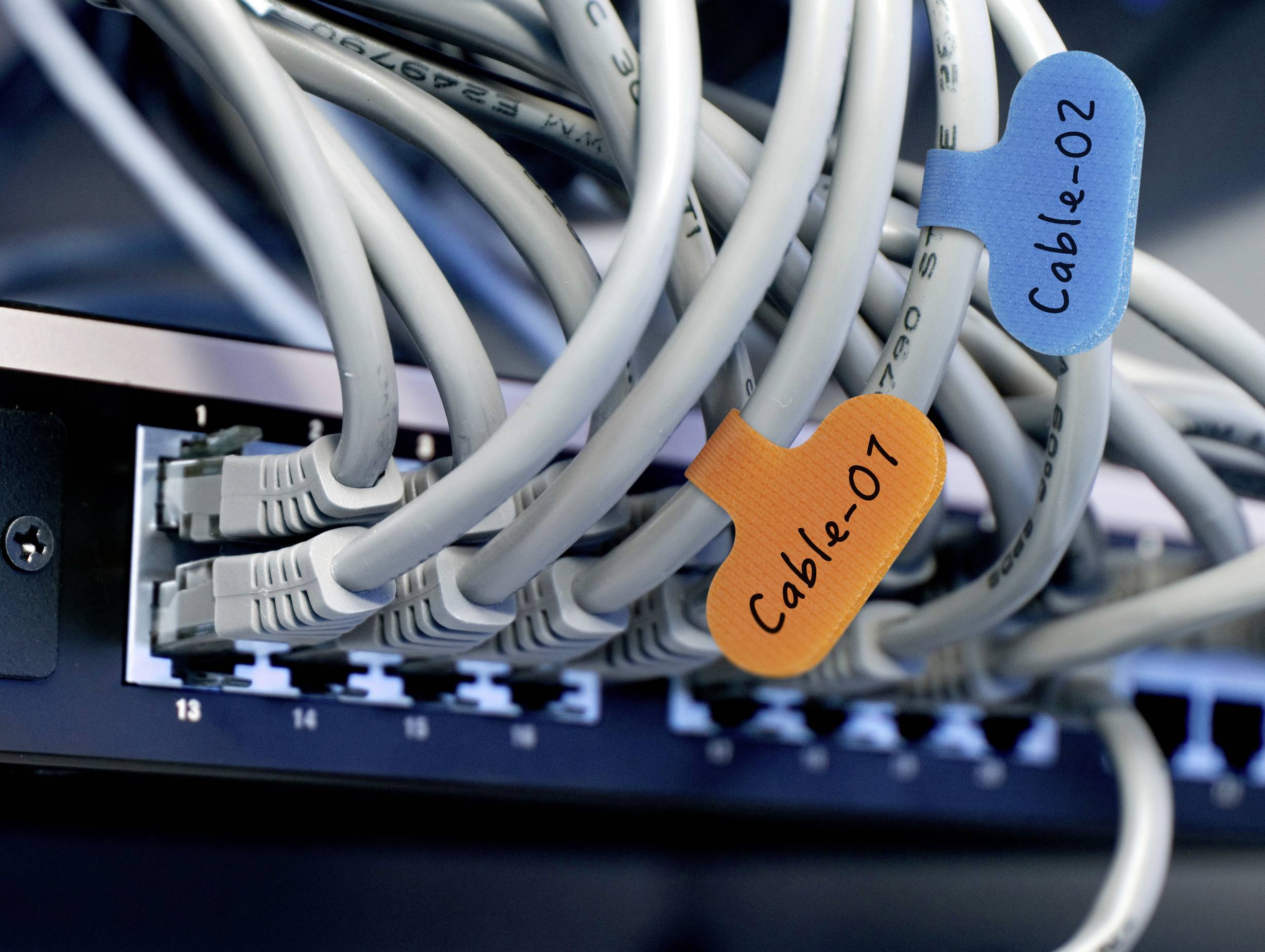 Nahaufnahme von Netzwerkkabeln, die an einen Switch angeschlossen sind, beschriftet mit 'Kabel-01' mit einem orangefarbenen Etikett und 'Kabel-02' mit einem blauen Etikett.