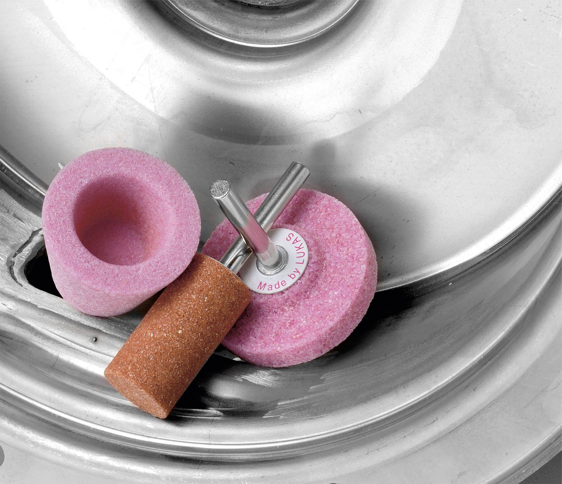 Polierwerkzeuge einschließlich eines rosa Schleifrades, eines braunen Schleifsteins und einer Polierspitze auf einer Metalloberfläche, was auf maschinelle Verwendung hindeutet.