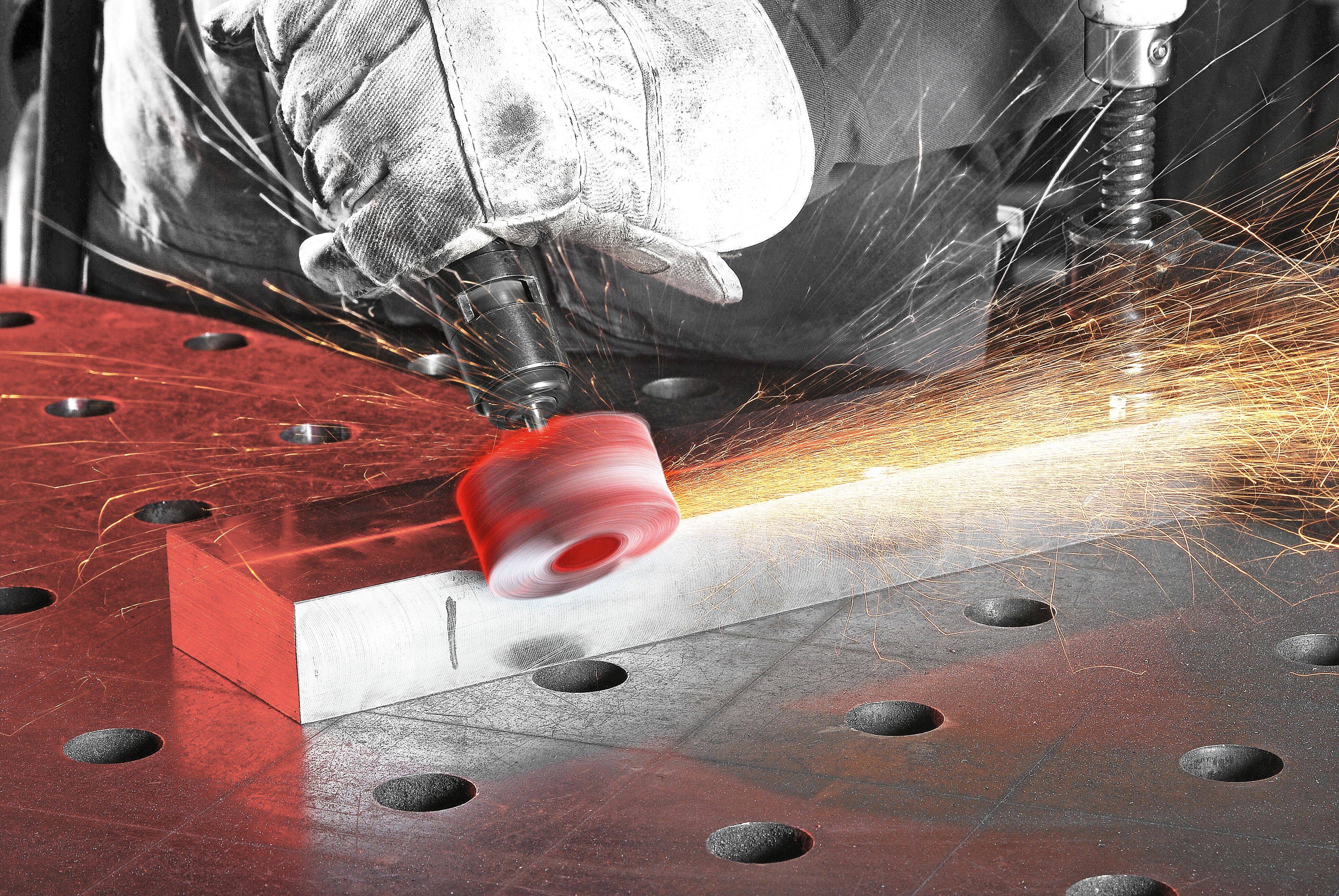 Ein Arbeiter schleift ein Metallstück auf einem gelochten Tisch und erzeugt dabei helle Funken. Der Arbeiter trägt Handschuhe, was den Fokus auf Sicherheit unterstreicht.