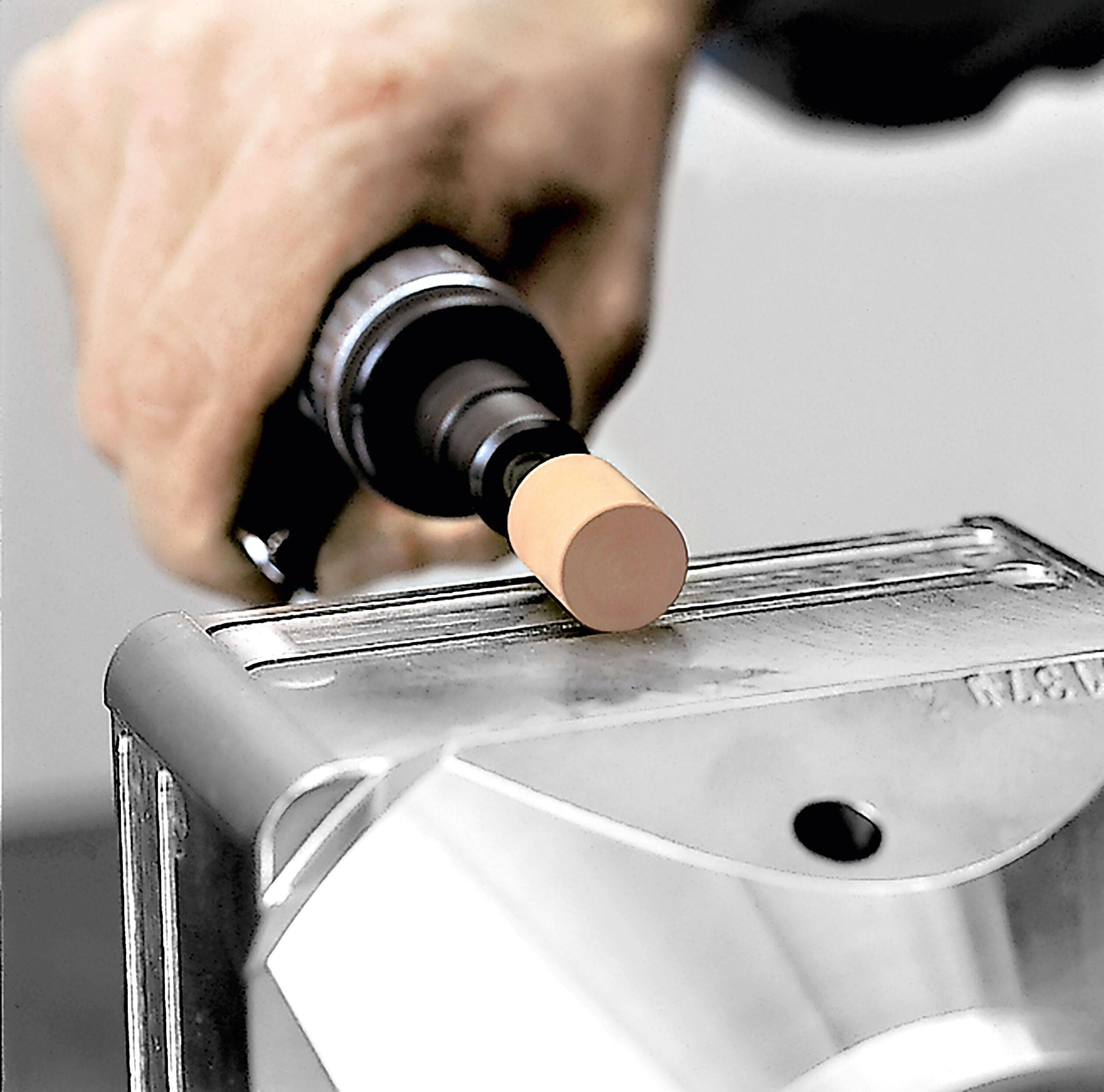 Eine Hand, die eine schwarze Flasche mit spezieller Flüssigkleber hält und diesen auf ein rechteckiges Objekt aufträgt, das in einem kleinen Metallschraubstock befestigt ist.