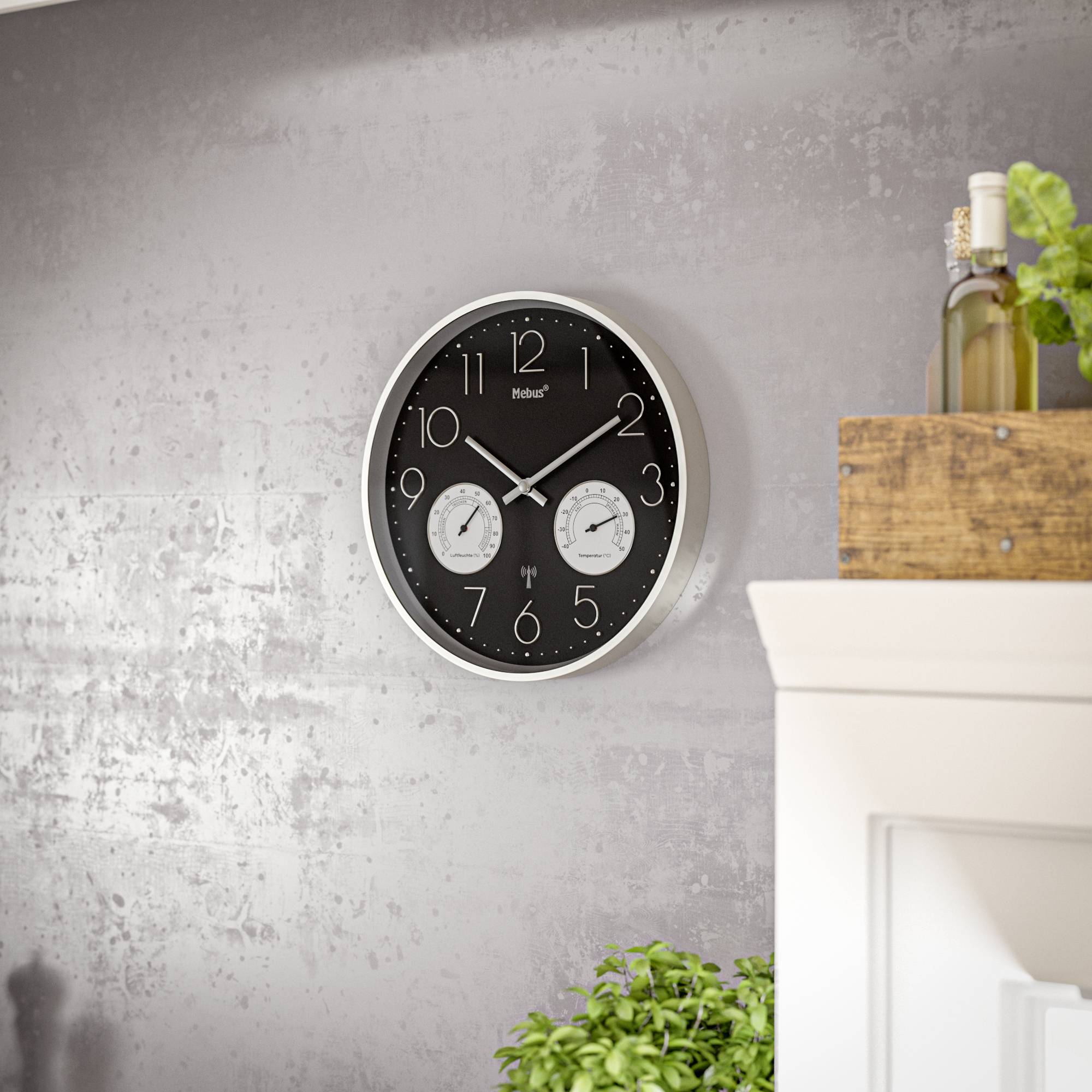 Eine runde Wanduhr mit zwei kleineren Zifferblättern für Temperatur und Luftfeuchtigkeit, an einer strukturierten grauen Wand montiert, mit einer grünen Pflanze darunter.