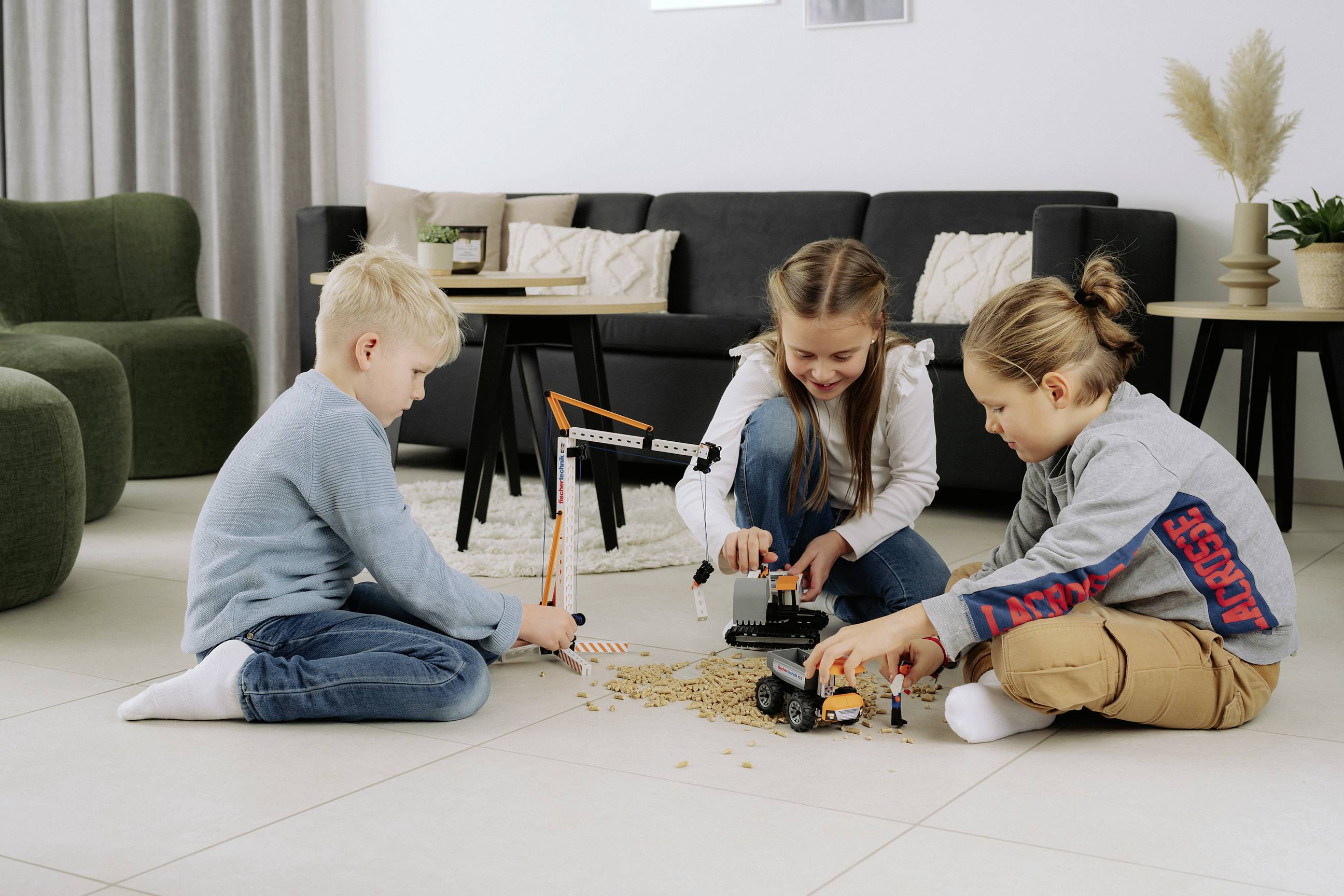 Drei Kinder spielen auf dem Boden mit Spielzeug-Baufahrzeugen und geben sich ihrer Fantasie hin. Im Hintergrund befindet sich ein schwarzes Sofa und einige Pflanzen.