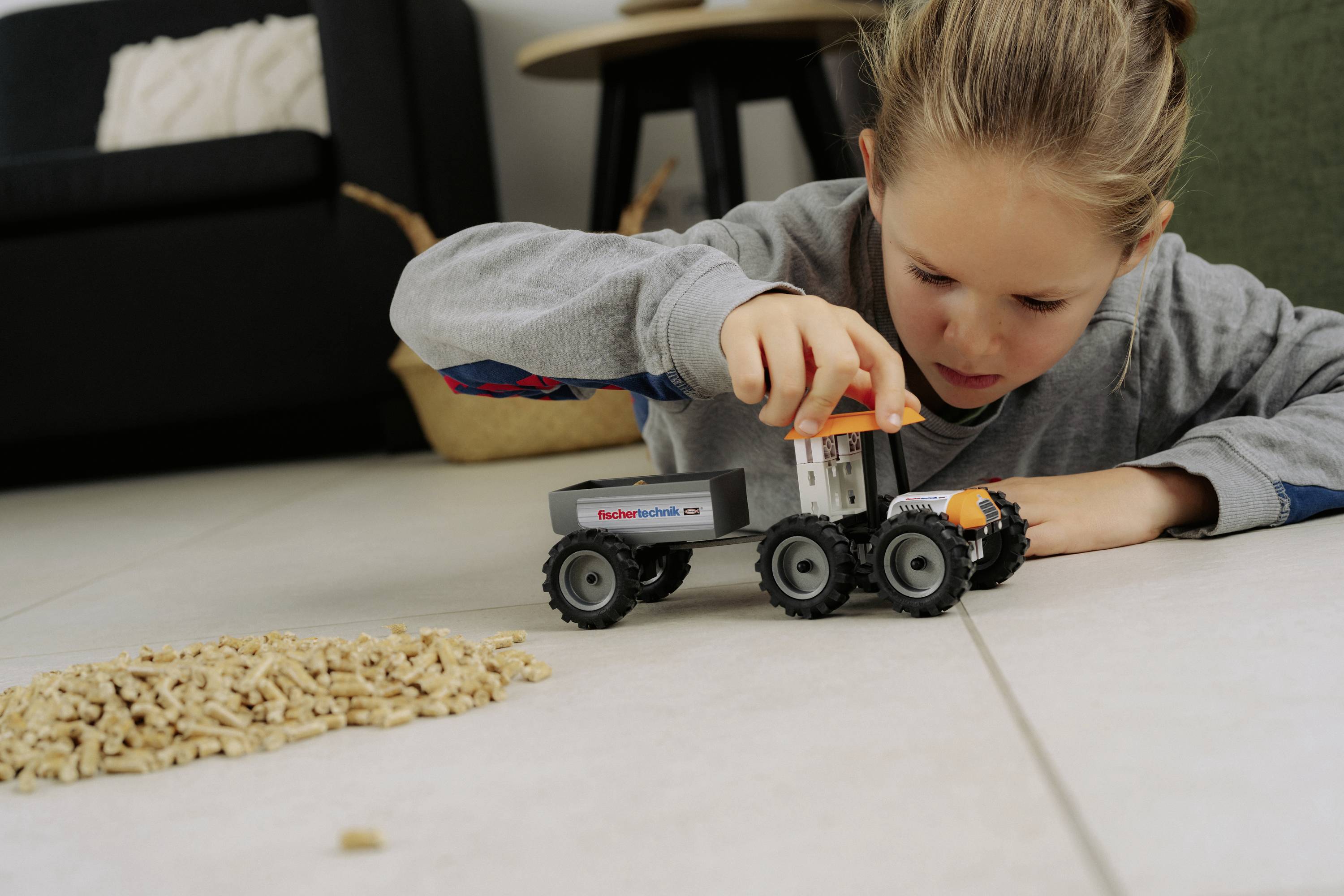 Ein kleines Kind, das in einem gemütlichen Wohnzimmer auf einem gefliesten Boden mit einem Spielzeugtraktor spielt, neben einem kleinen Haufen Körner.