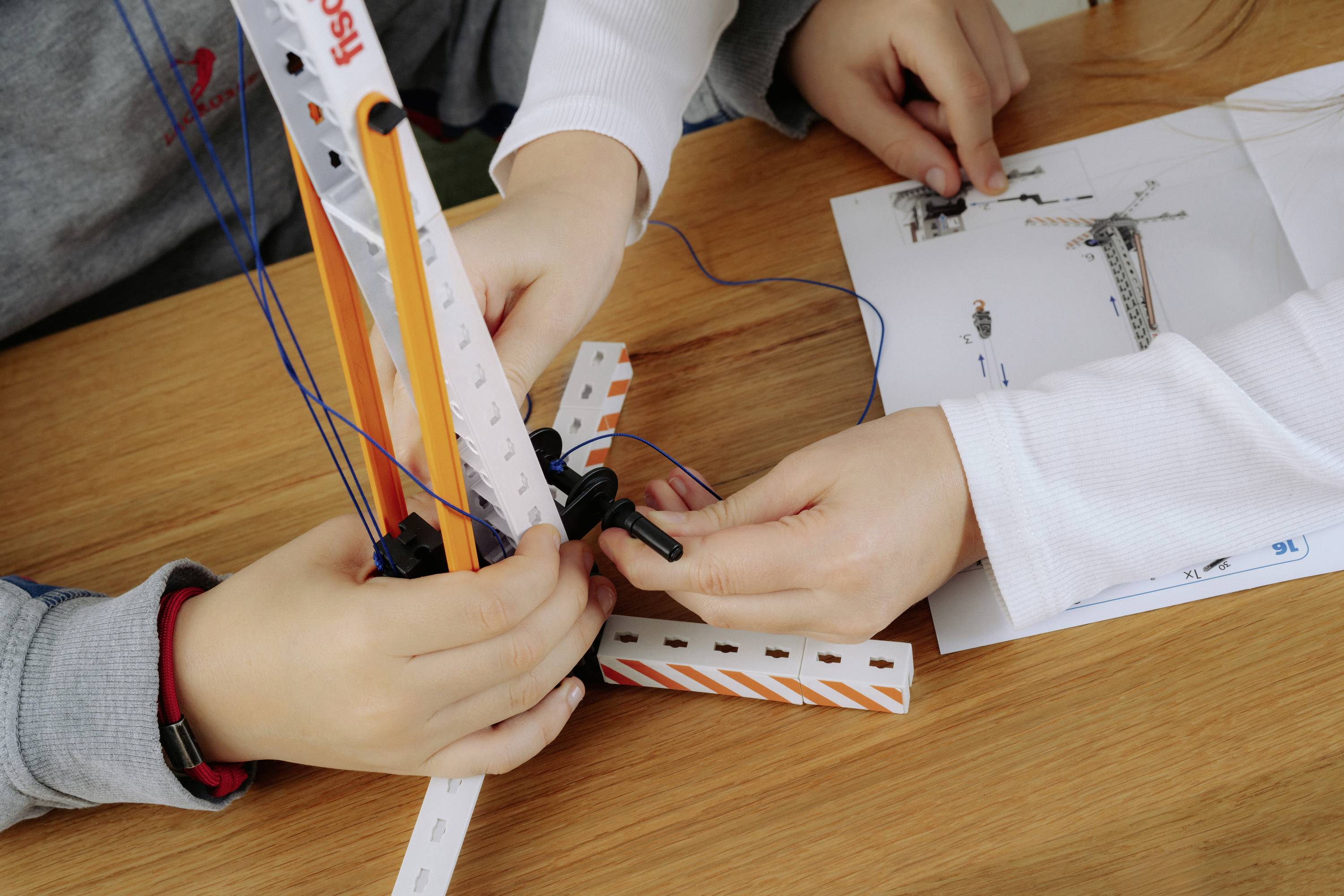 Zwei Kinder bauen eine Modellstruktur mit Plastikstäben und Verbindungselementen. Ein gedrucktes Anleitungsblatt ist teilweise auf dem Holztisch zu sehen.