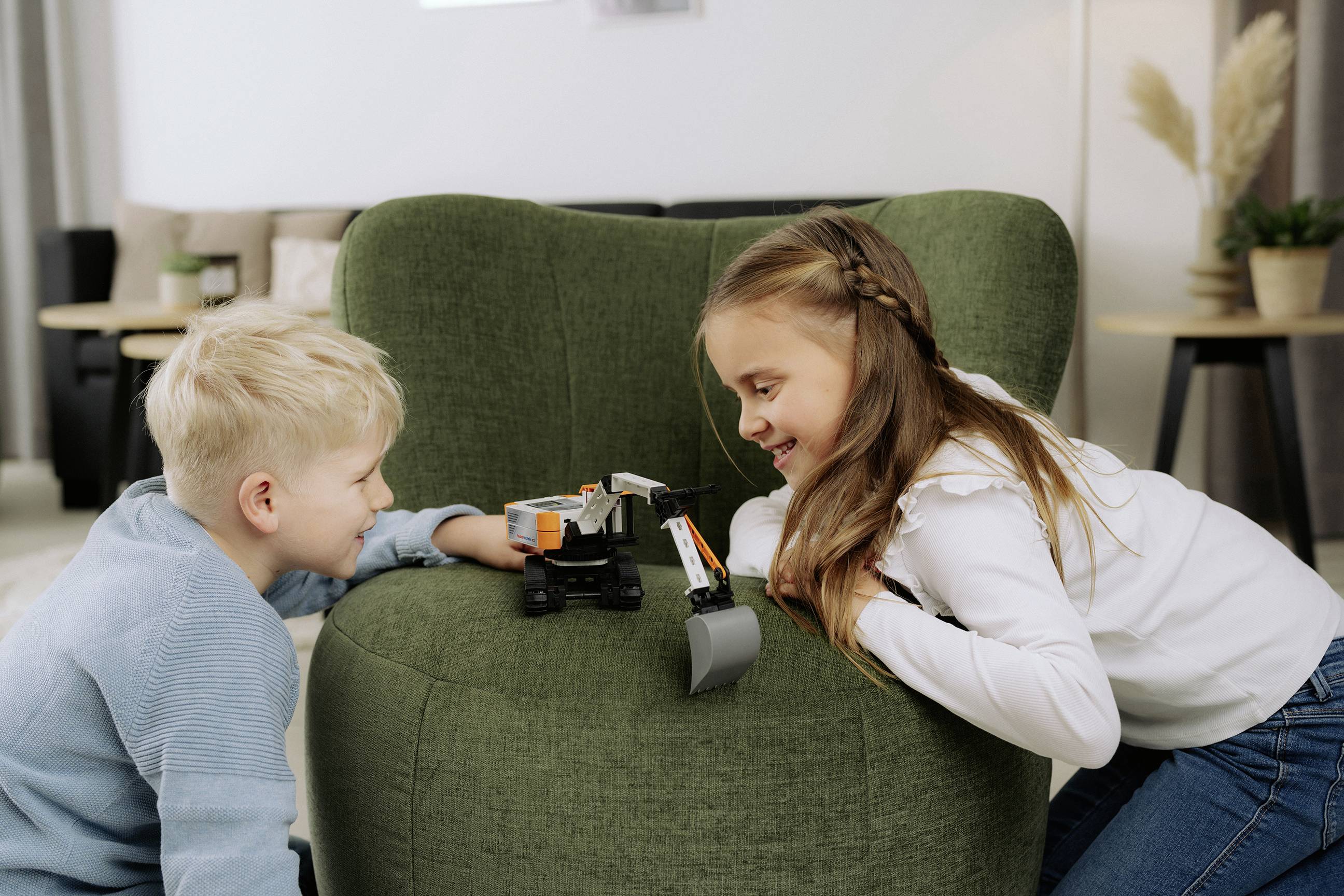 Zwei Kinder lächeln und spielen mit einem Spielzeug-Bulldozer auf einem grünen Stuhl in einer gemütlichen Wohnzimmeratmosphäre.