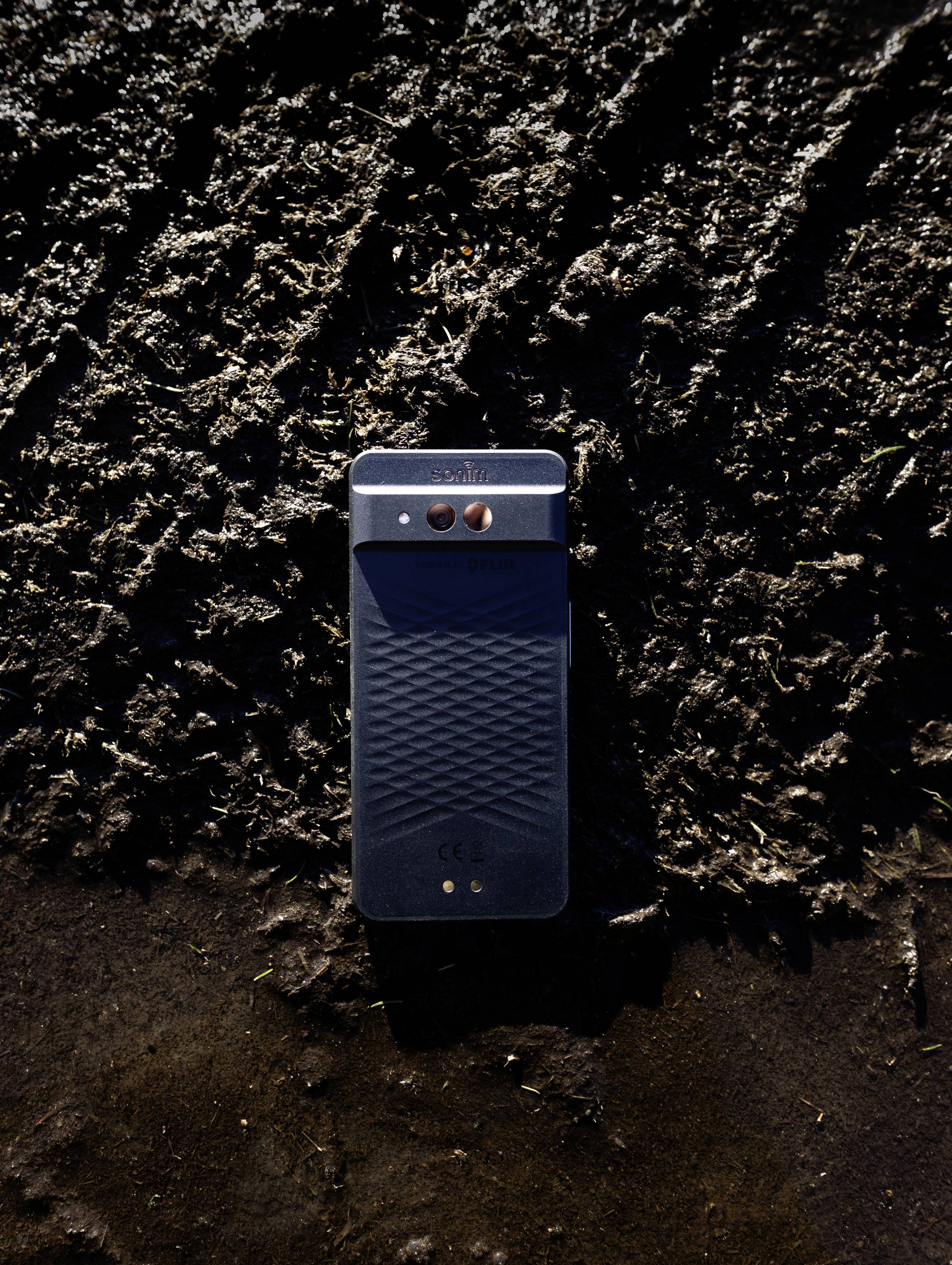 Ein Smartphone mit strukturierter Rückseite liegt auf dunklem, schlammigem Untergrund und demonstriert sein robustes Design und seine Widerstandsfähigkeit in rauen Umgebungen.