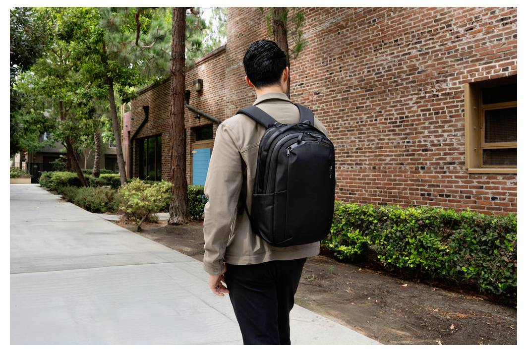 Eine Person, die sich von einem ziegelbaugebäude entfernt, geht an einem baumbestandenen Gehweg vorbei und trägt einen großen schwarzen Rucksack.