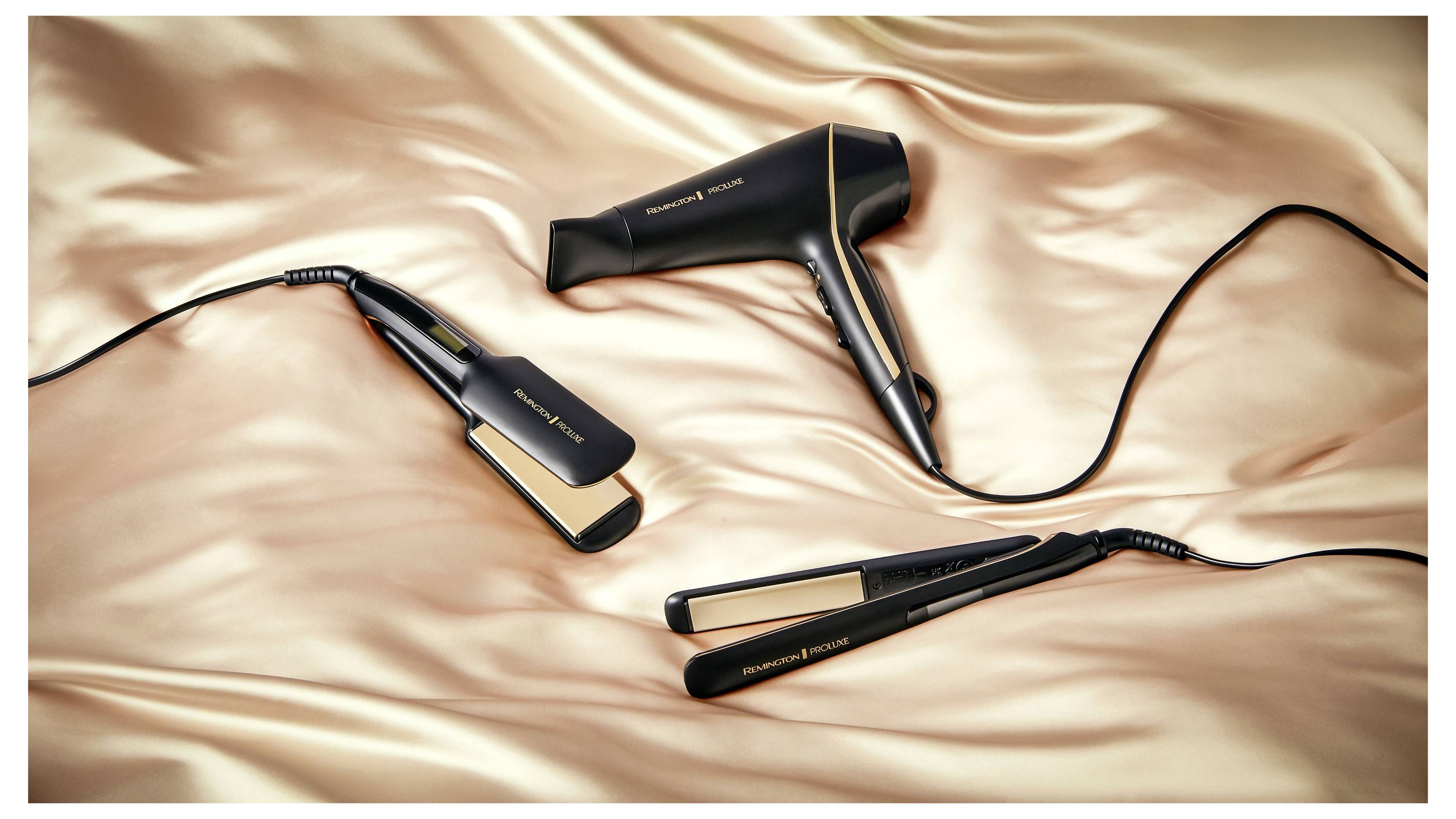 Schwarze und goldene Haarstyling-Geräte, darunter ein Haarföhn und zwei Glätteisen, vor einem seidigen Satin-Hintergrund.