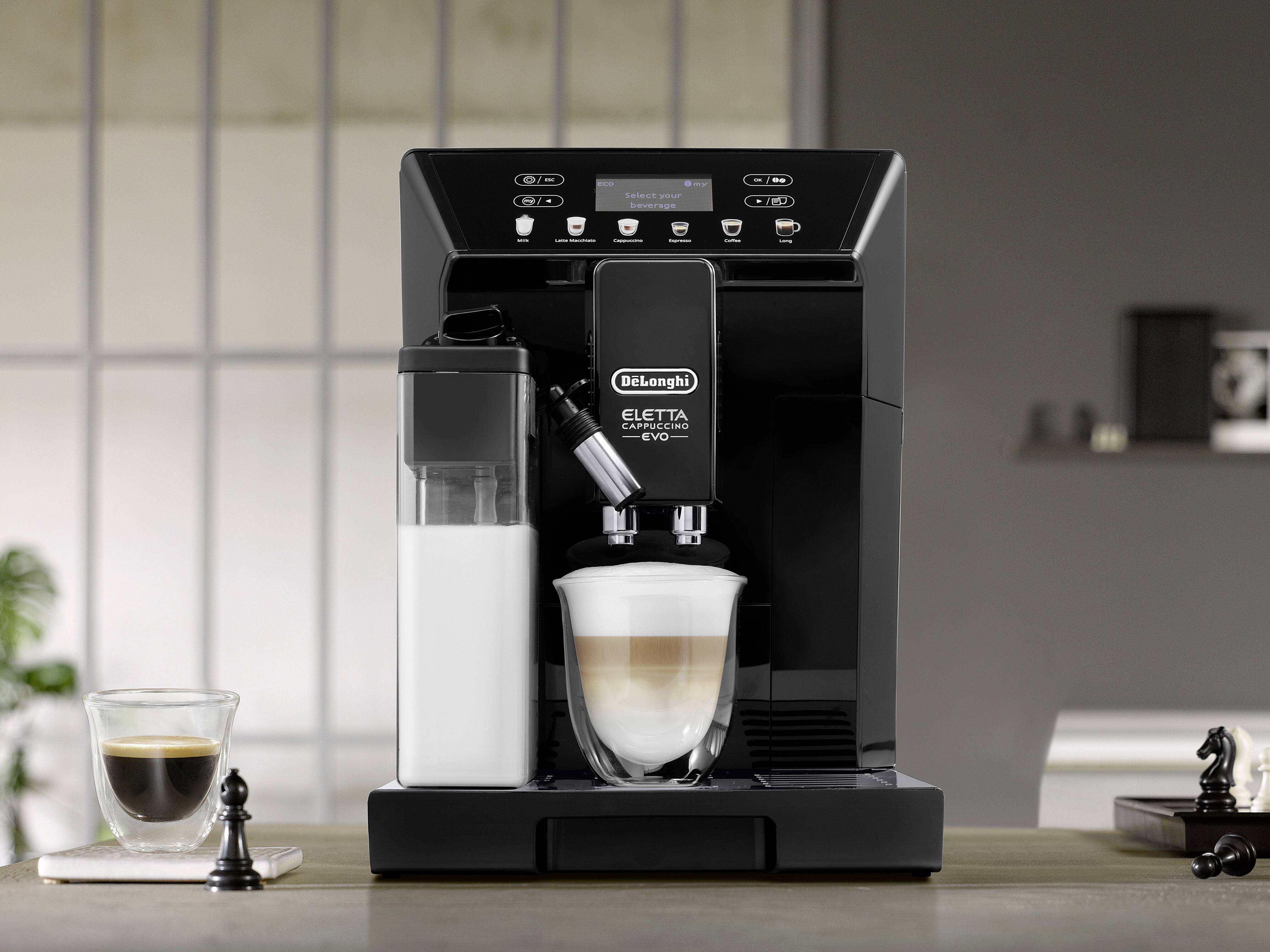 Eine schwarze Espressomaschine mit einem Milchaufschäumer bereitet einen Cappuccino in einem Glas zu. In der Nähe stehen eine Tasse Espresso und Schachfiguren.