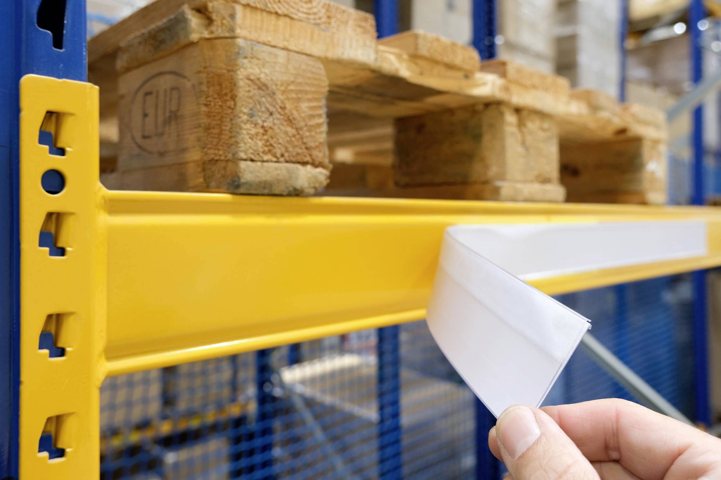Eine Hand befestigt ein leeres Etikett an einem gelben Lagerregal, mit Holzpaletten darüber, was auf Organisation oder Bestandsmanagement hindeutet.