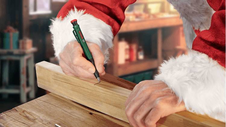 Eine Person in einem Weihnachtsmannkostüm, die ein Holzbrett mit einem Bleistift auf einer Werkbank vermisst, umgeben von einer Werkstattumgebung.