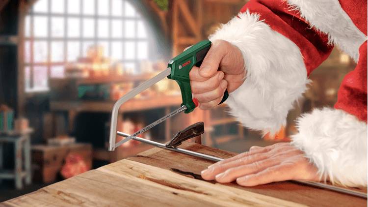 Eine Person, gekleidet in festliche rot-weiße Kleidung, schneidet mit einer kleinen Säge ein Stück Holz in einer Werkstatt, was auf Feiertagsvorbereitungen hindeutet.