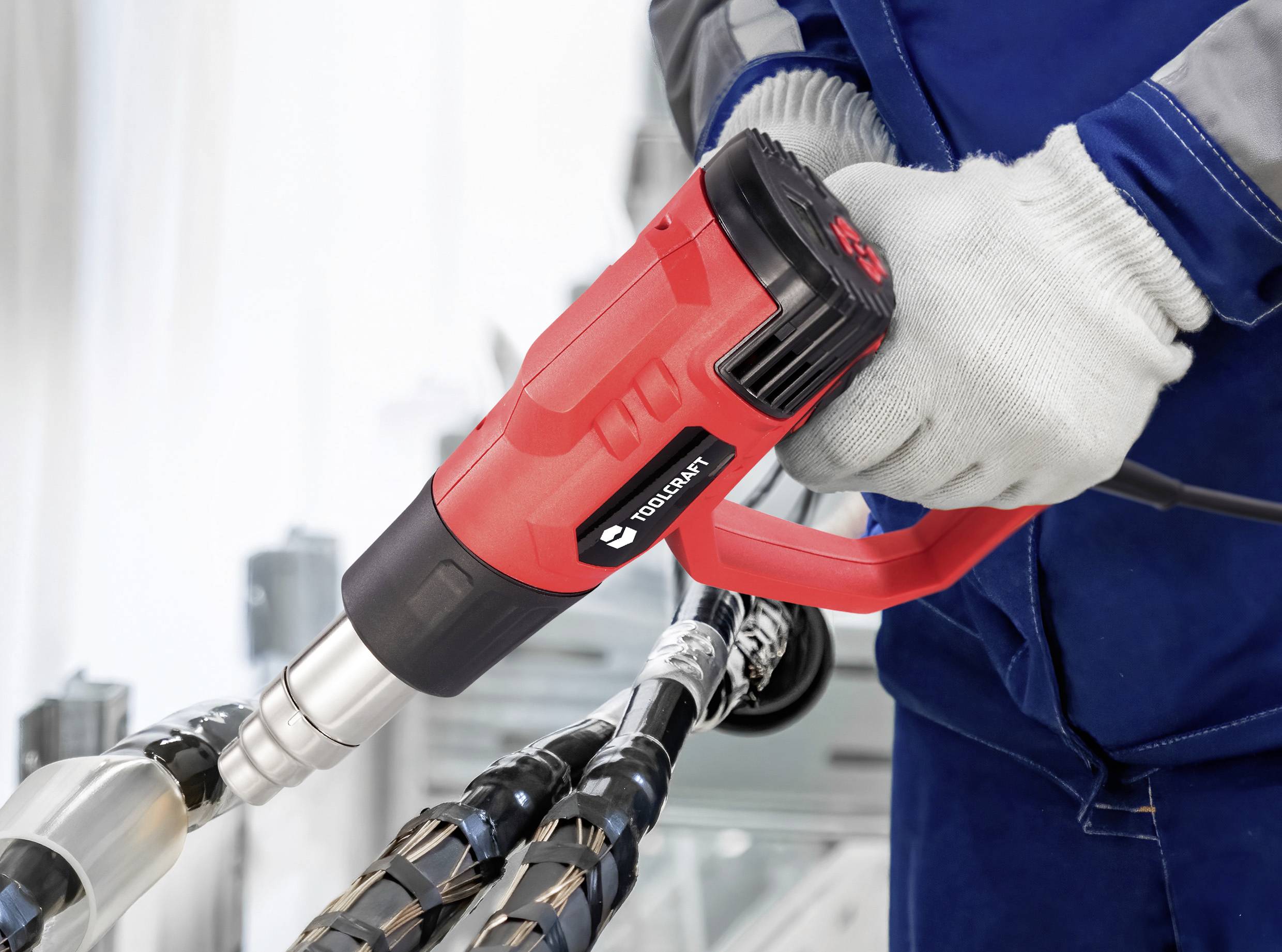 Eine Person in blauem Arbeitsanzug und Handschuhen verwendet eine rote Heißluftpistole auf schwarzen Kabeln, was auf eine technische oder Reparaturaufgabe in einer professionellen Umgebung hindeutet.