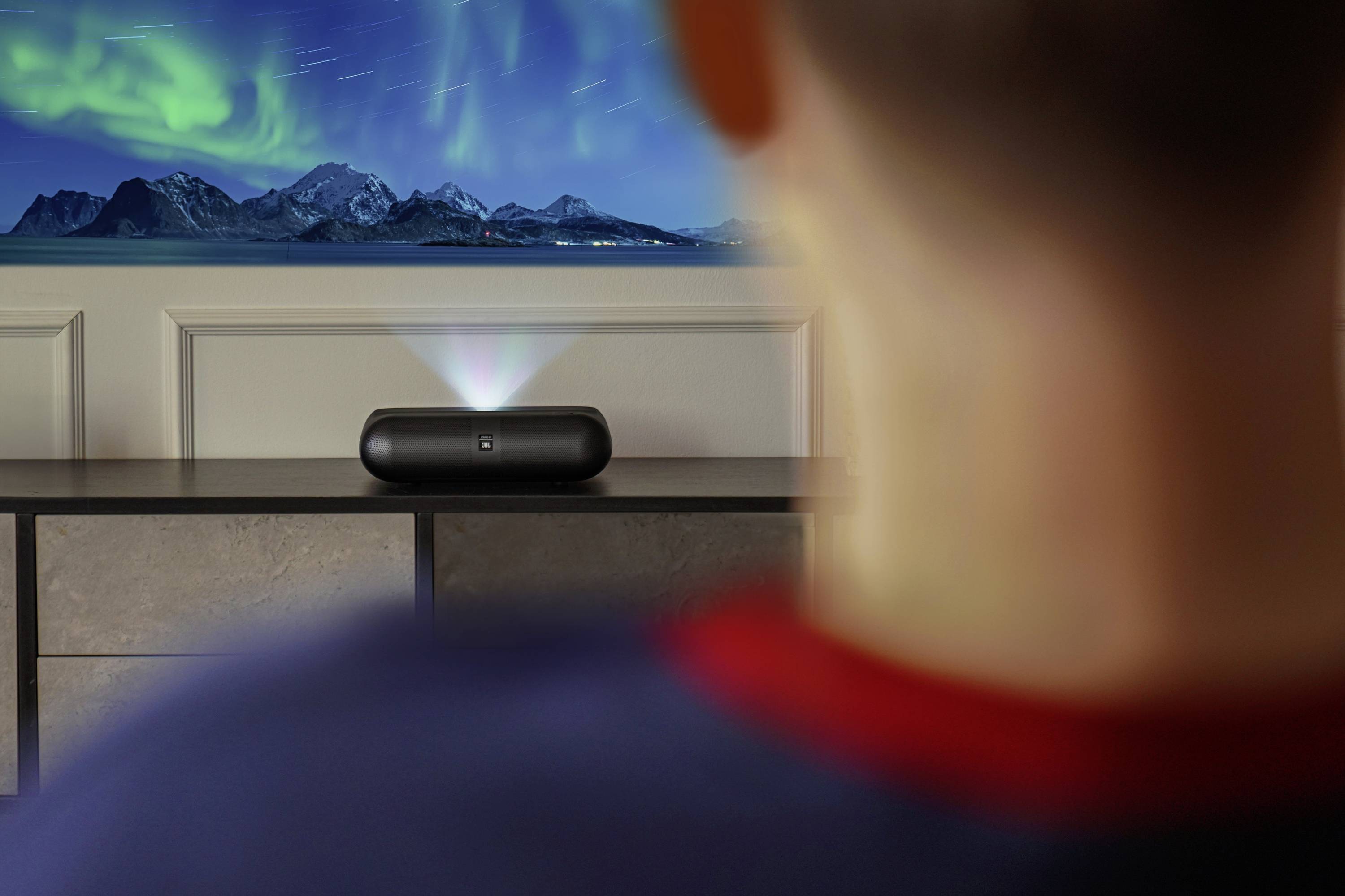 Eine Person beobachtet ein Nordlicht, das von einem schwarzen, lautsprecherähnlichen Gerät auf einem Schrank an die Wand projiziert wird. Berge sind in der Projektion zu sehen.