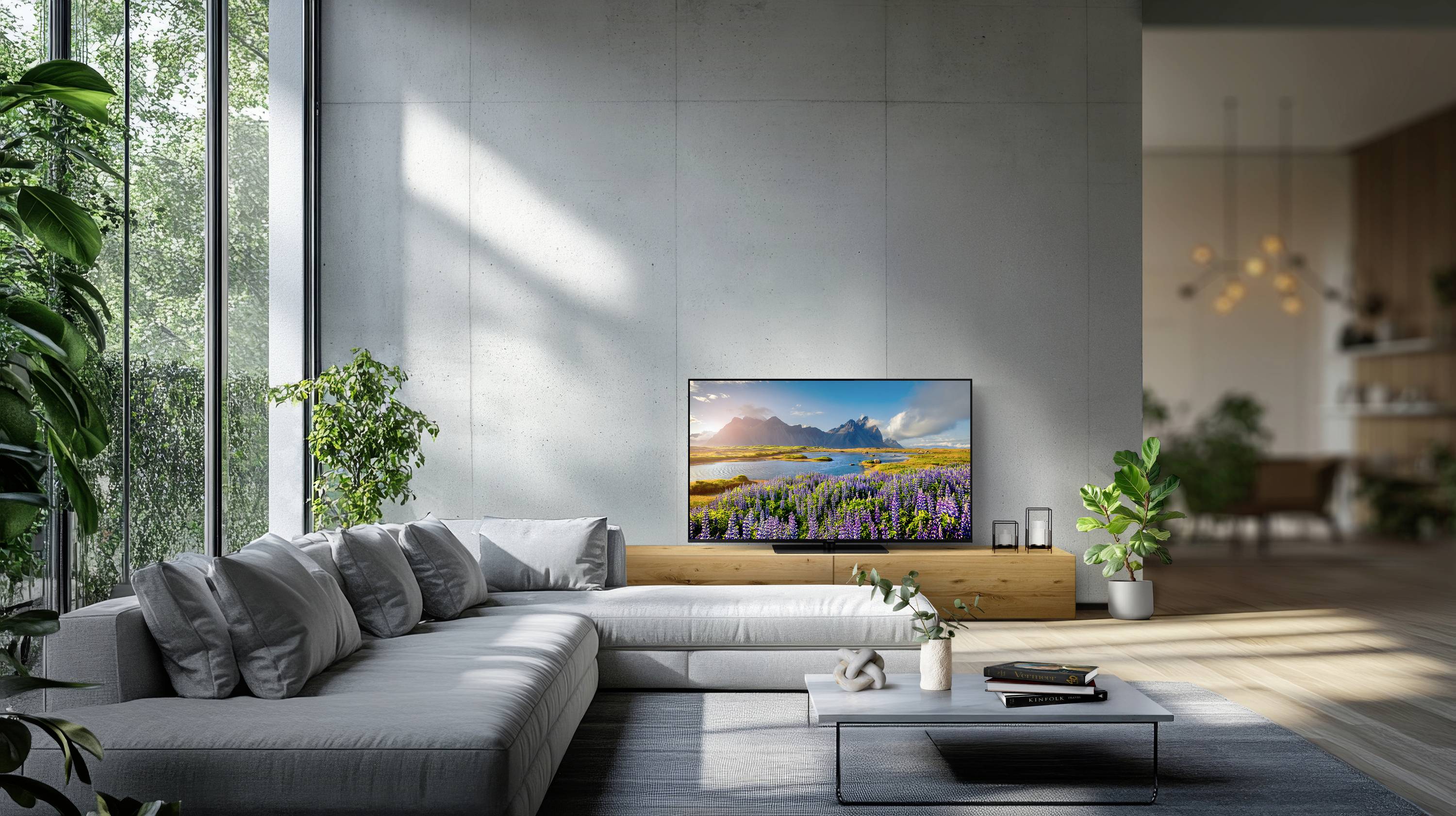 Ein modernes Wohnzimmer mit einem großen grauen Ecksofa, einem an der Wand montierten Fernseher, der eine malerische Landschaft zeigt, und Pflanzen neben großen Fenstern.
