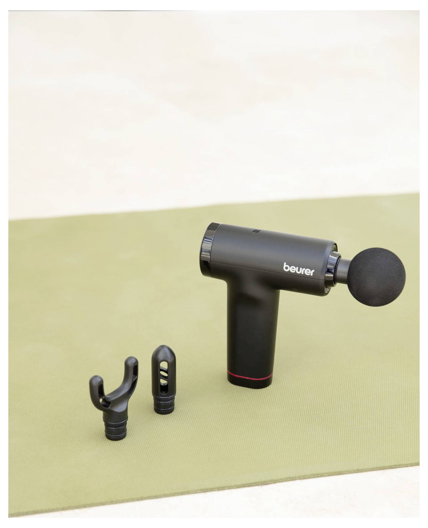 Eine schwarze, handgehaltene Massagepistole mit drei abnehmbaren Aufsätzen liegt auf einer beigen Oberfläche, zur Produktwerbung präsentiert.