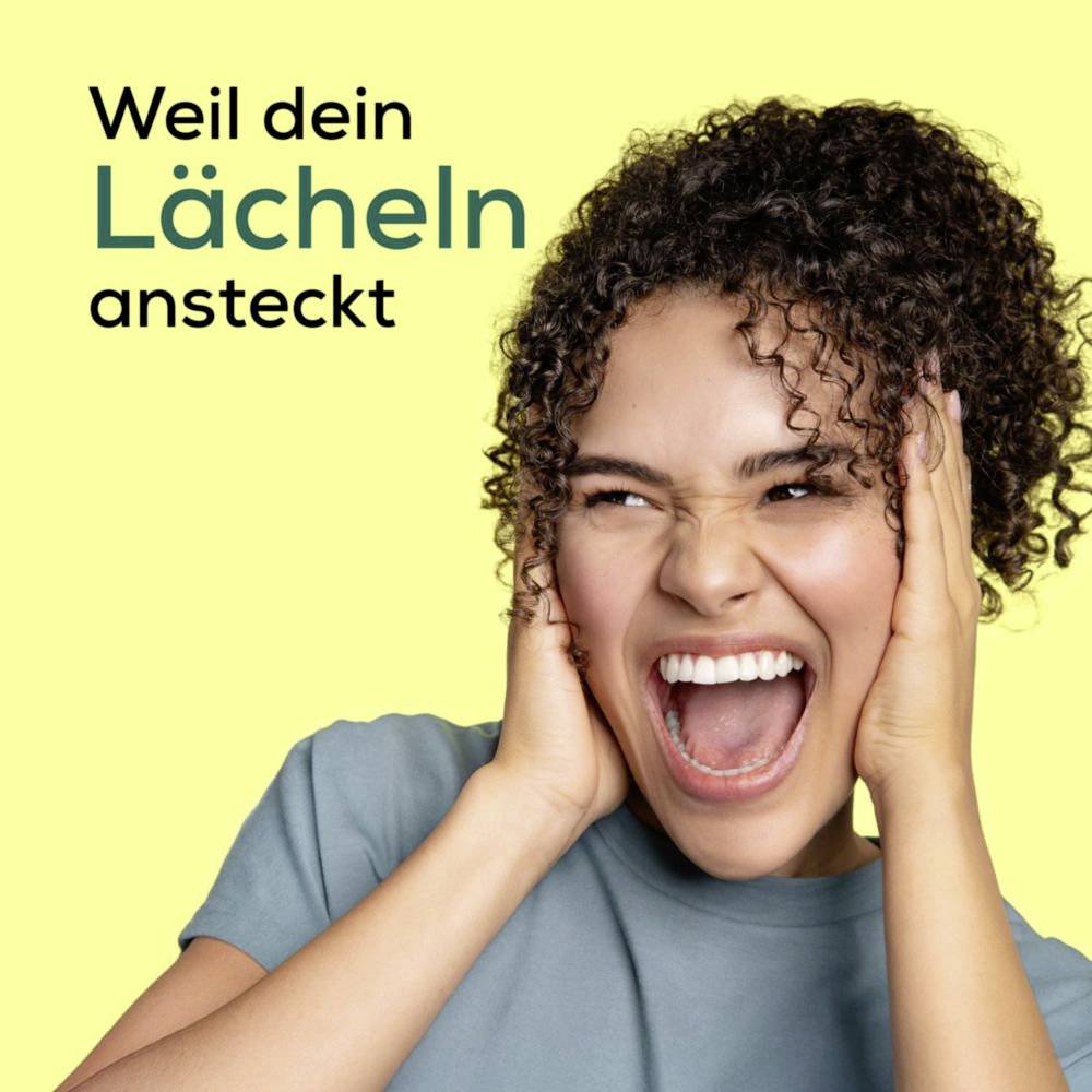Eine Frau mit lockigem Haar lächelt breit und hält sich vor Aufregung das Gesicht. Der Text lautet "Weil dein Lächeln ansteckt" auf einem gelben Hintergrund.