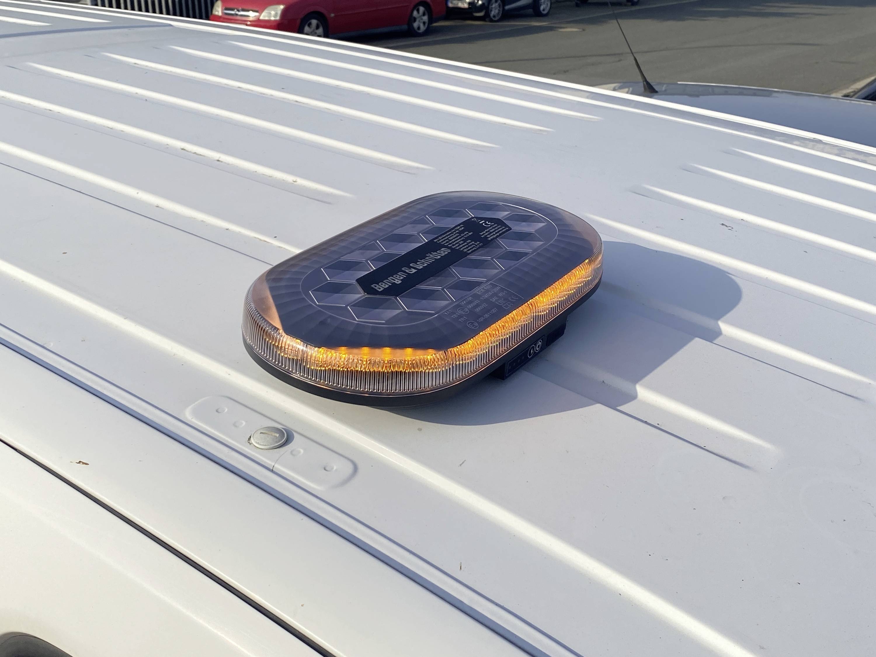 Eine solarbetriebene Bernsteinfarben-Warnleuchte, die auf dem weißen Dach eines Fahrzeugs montiert ist und leuchtet, mit mehreren Autos im Hintergrund auf einem Parkplatz.