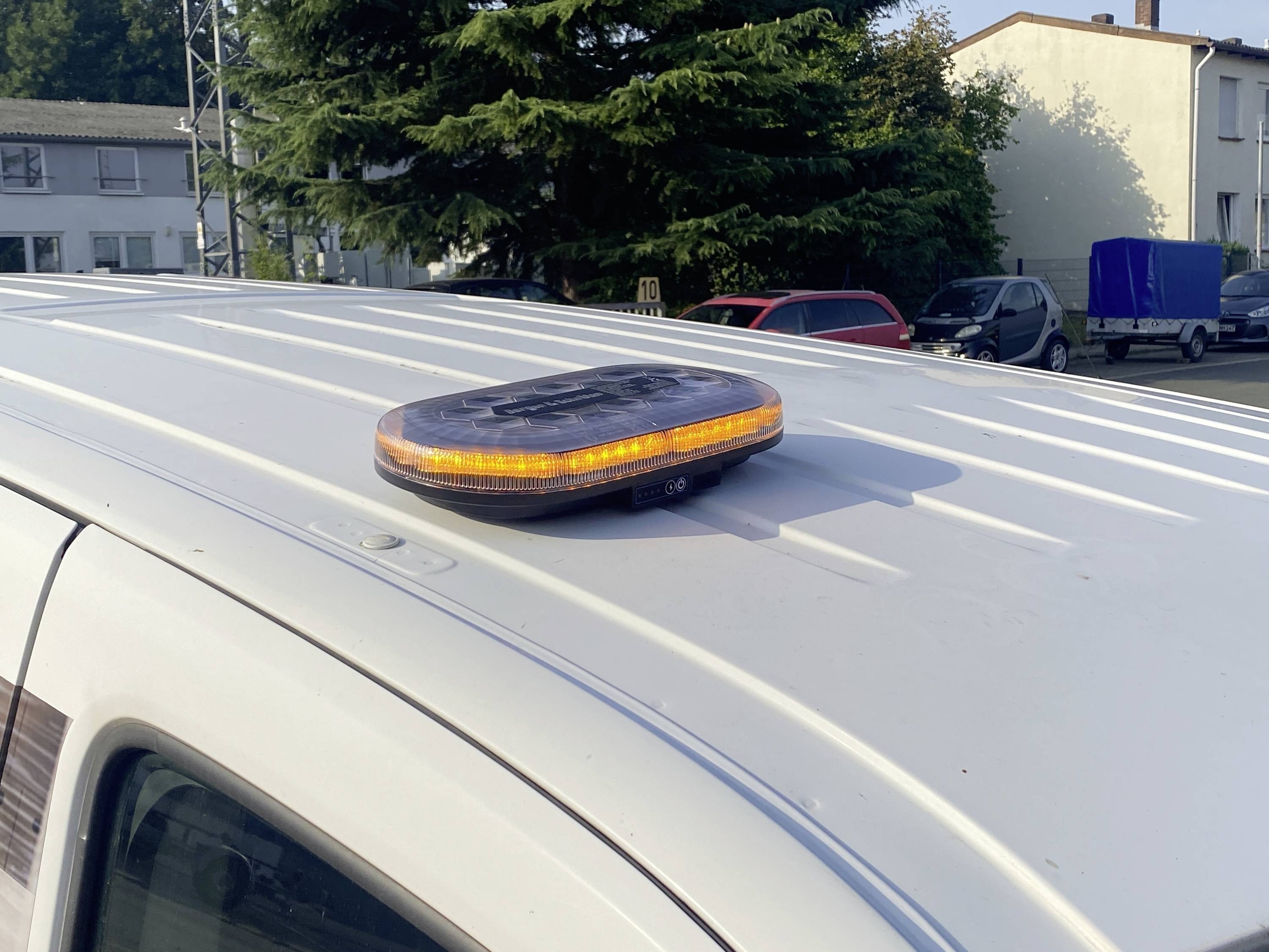 Eine Dachaufbau-Warnleuchte in Bernsteinfarbe auf einem weißen Fahrzeug, geparkt in der Nähe von Gebäuden und Bäumen, die ihre Verwendung für Sichtbarkeit oder Vorsicht hervorhebt.