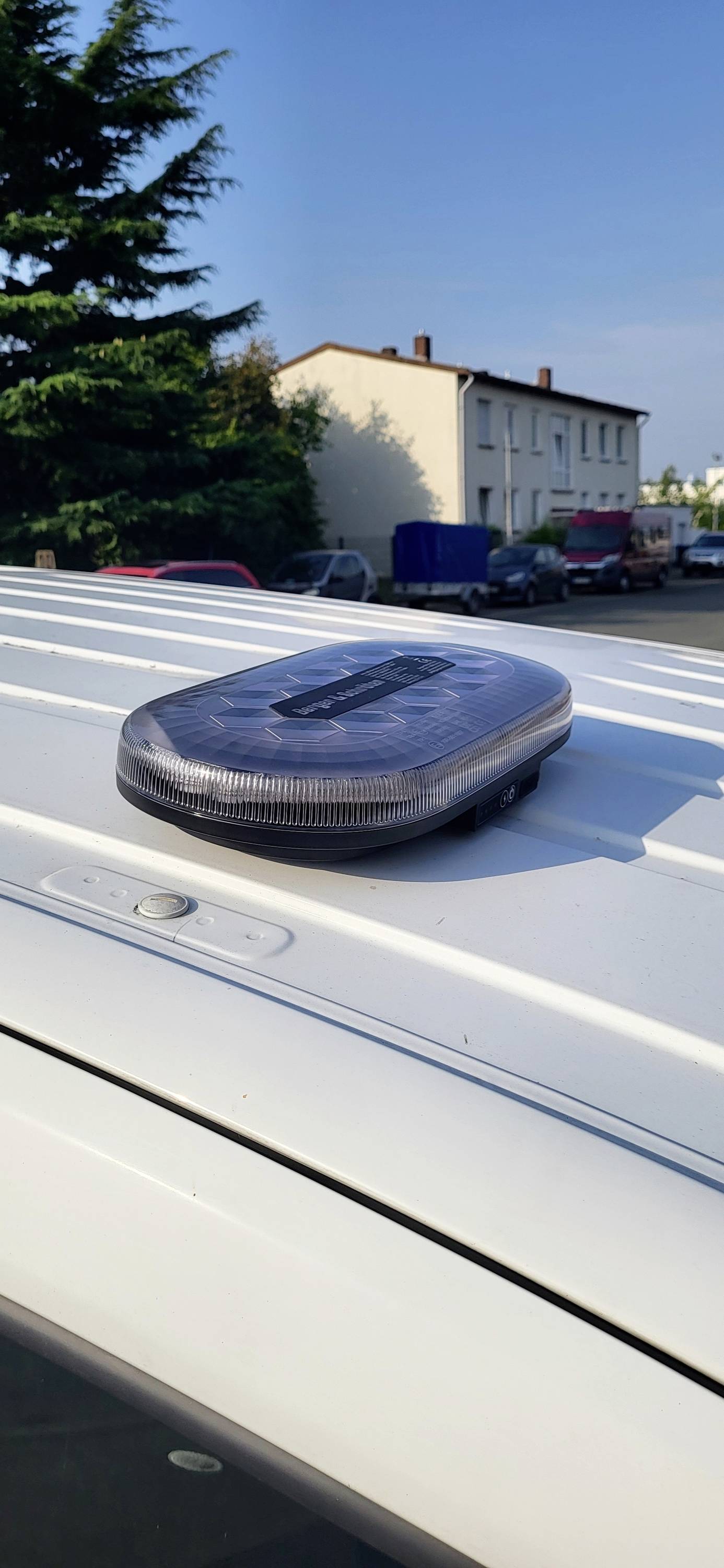 Ein Solarpanel ist auf dem Dach eines weißen Fahrzeugs montiert, das im Freien geparkt ist, mit Bäumen und Gebäuden im Hintergrund.