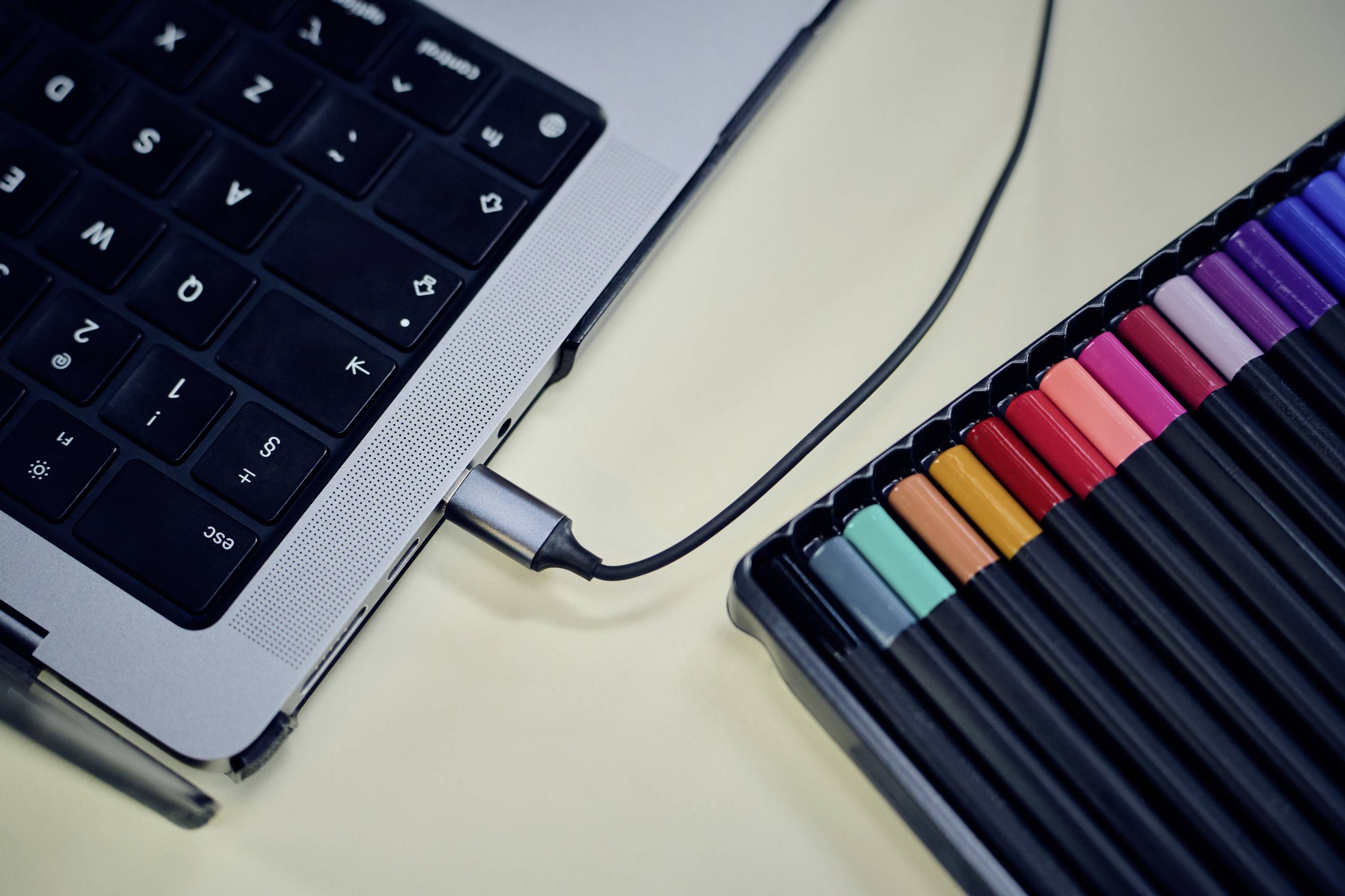 Ein geschlossener Laptop mit einem angeschlossenen Kabel liegt neben einem Satz bunter Marker und deutet auf Kreativität und Arbeitsumgebung hin.