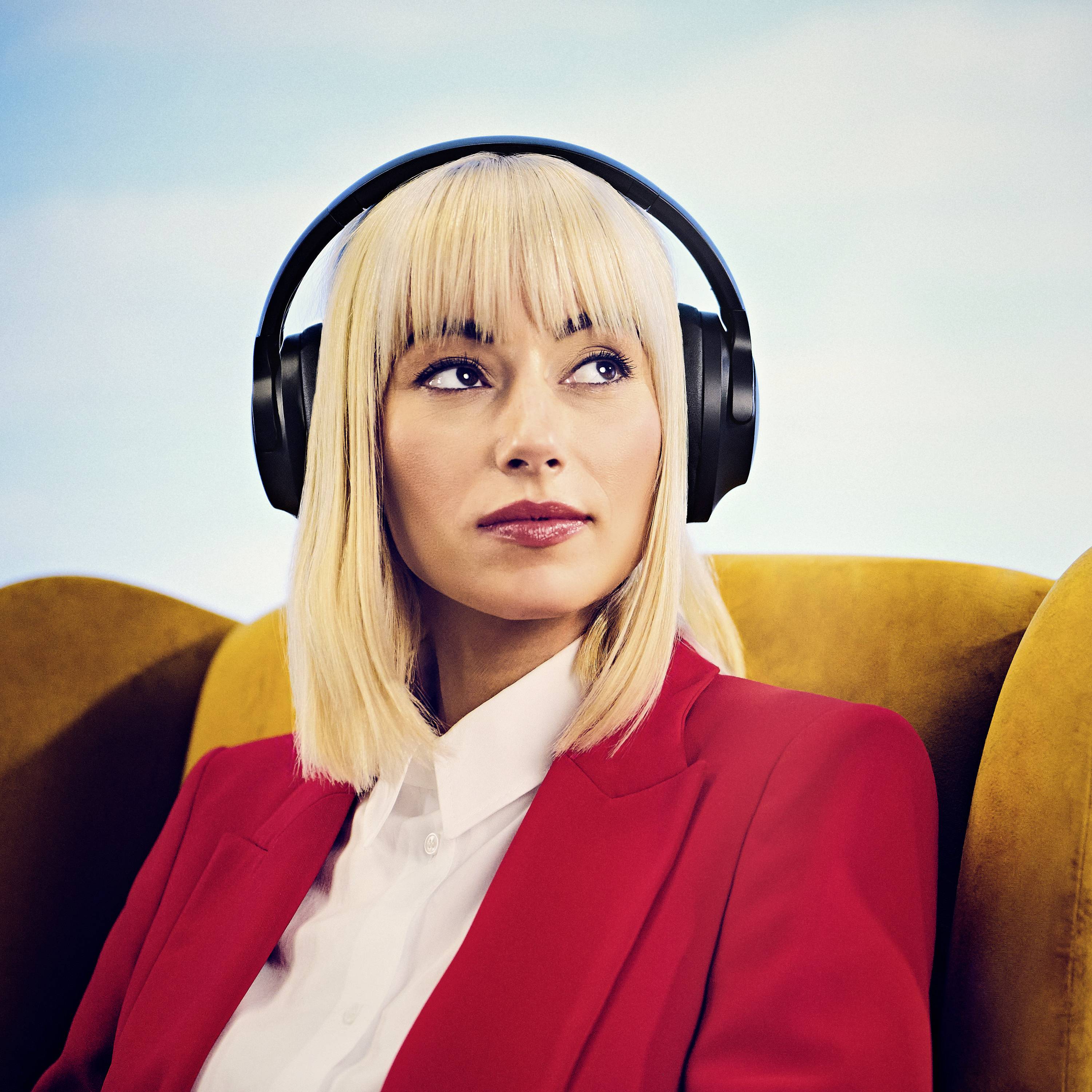 Eine Person mit Kopfhörern, in einem Stuhl sitzend, nachdenklich in die Ferne blickend. Sie trägt einen roten Blazer.