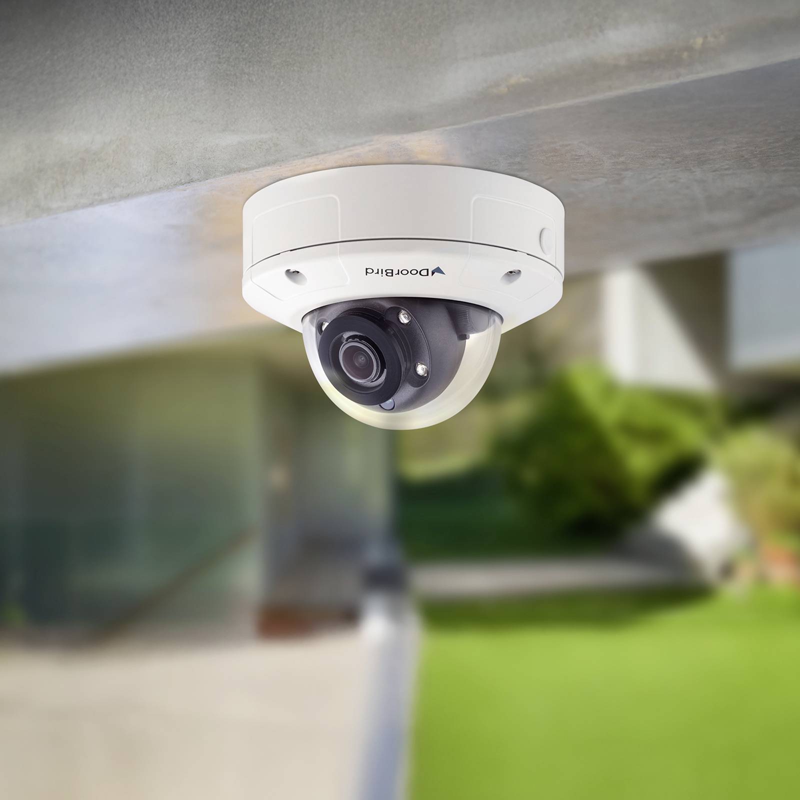 Sicherheitskamera an einer Decke im Freien montiert, mit Blick auf einen unscharf wirkenden grünen Rasen und ein modernes Gebäude im Hintergrund.