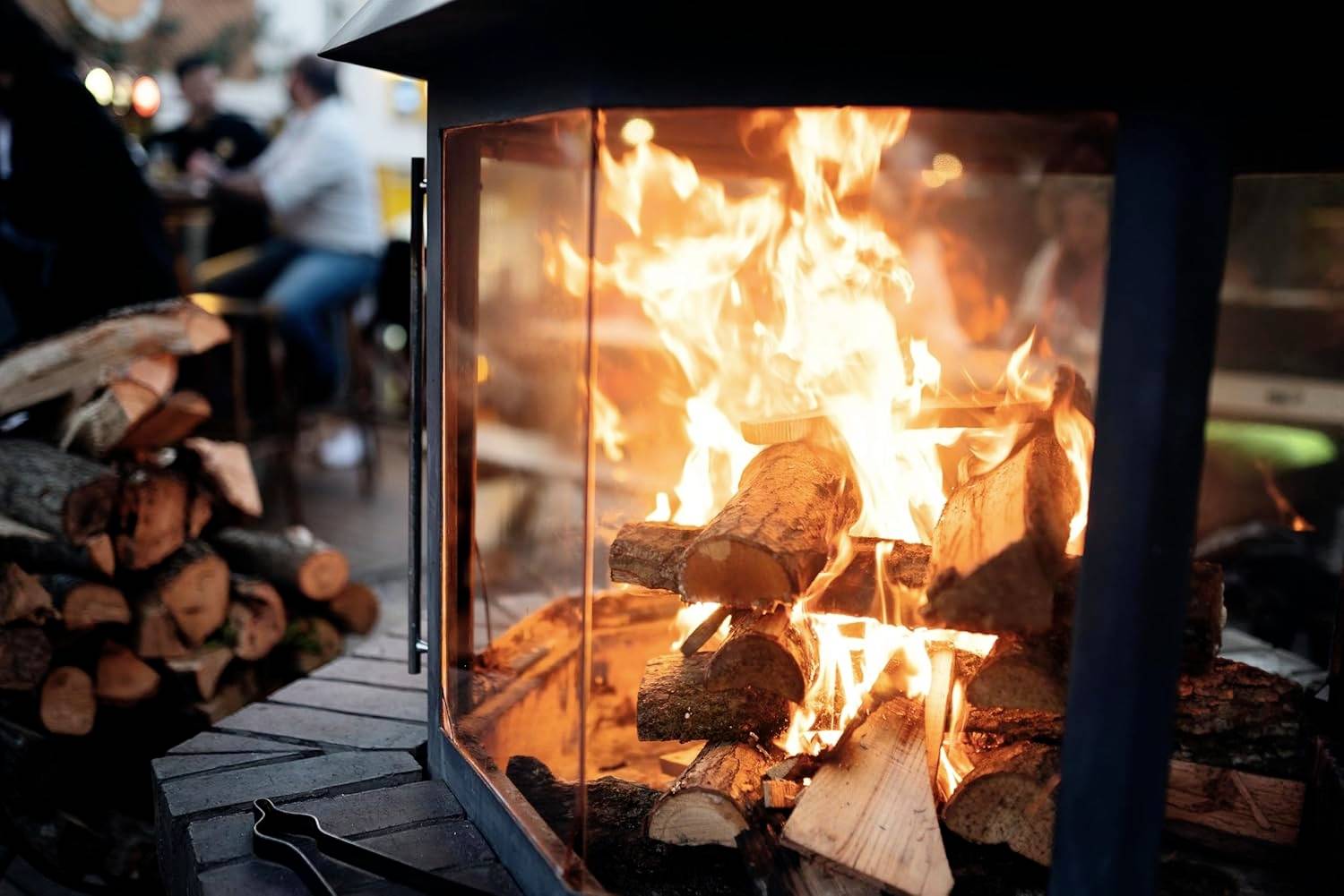 Ein gemütlicher Außenkamin mit lodernden Scheiten in einer Glasumhüllung, umgeben von einem Stapel Holz. Im Hintergrund sitzen Menschen und schaffen eine warme Atmosphäre.