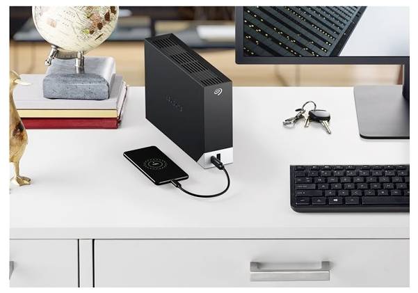Eine externe Festplatte liegt auf einem Schreibtisch und ist über ein Kabel mit einem Smartphone verbunden. Ein Monitor, eine Tastatur, ein Globus, Bücher und Schlüssel befinden sich in der Nähe.