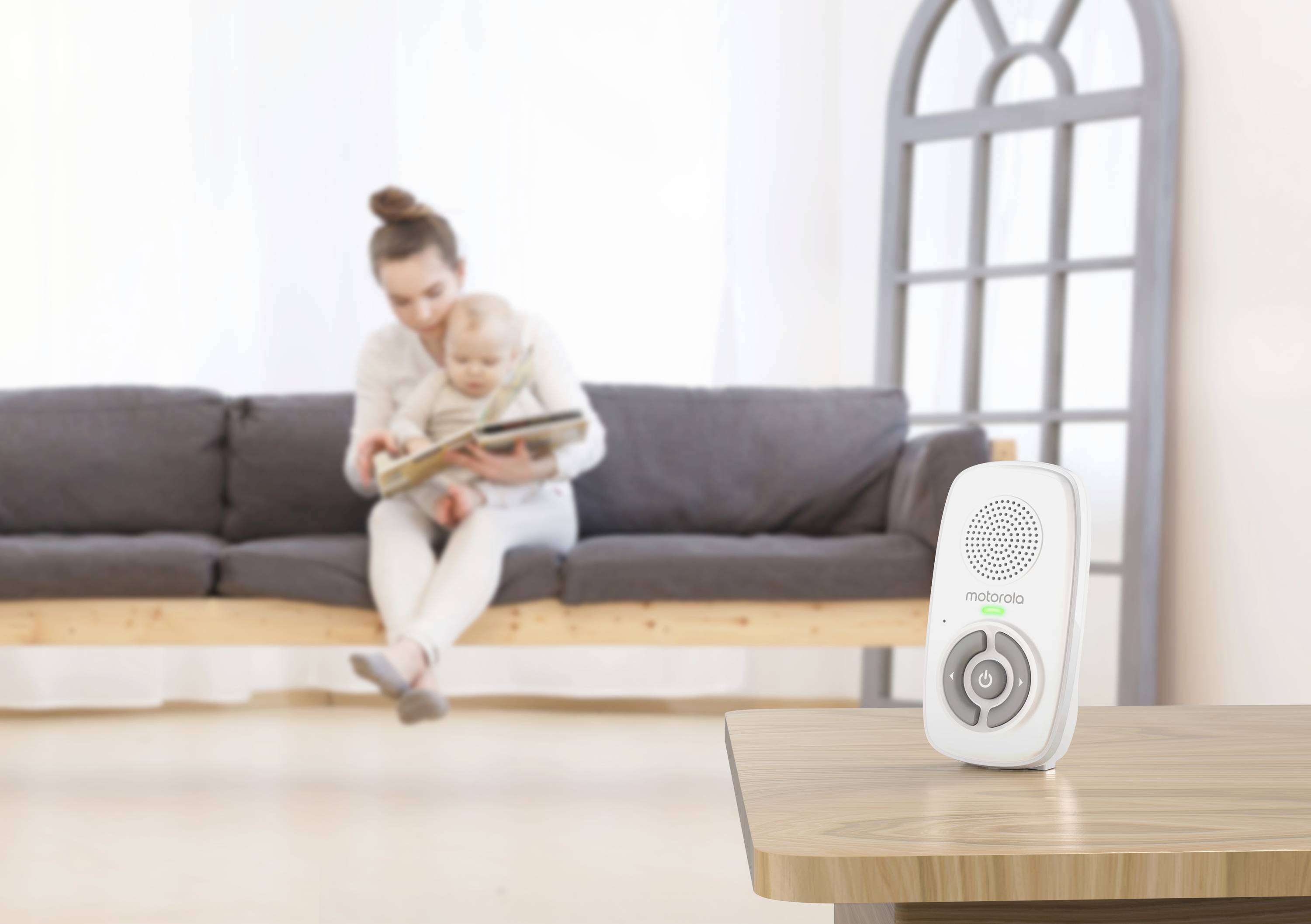 Ein Babyphone auf einem Holztisch im Vordergrund, während im unscharfen Hintergrund eine Frau einem Kind vorliest.